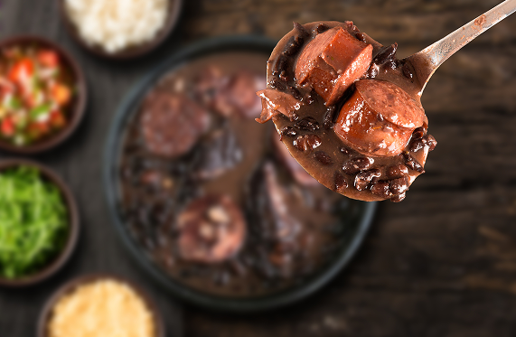 Uma colherada de feijoada brasileira com linguiça, servida sobre uma panela com acompanhamentos em uma mesa de madeira escura