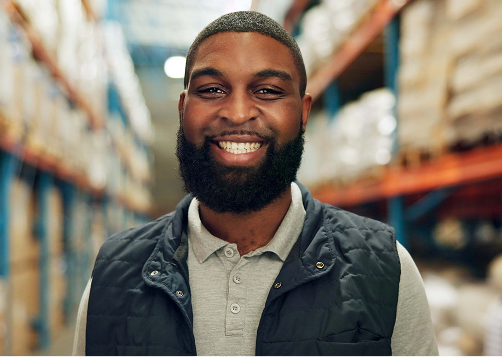 Homem sorrindo em um armazém, vestindo colete e camisa cinza, com prateleiras de mercadorias ao fundo.