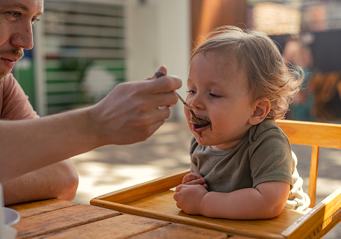 Pessoa alimentando um bebê em uma cadeirinha de alimentação em uma mesa ao ar livre. O bebê está com comida no rosto.