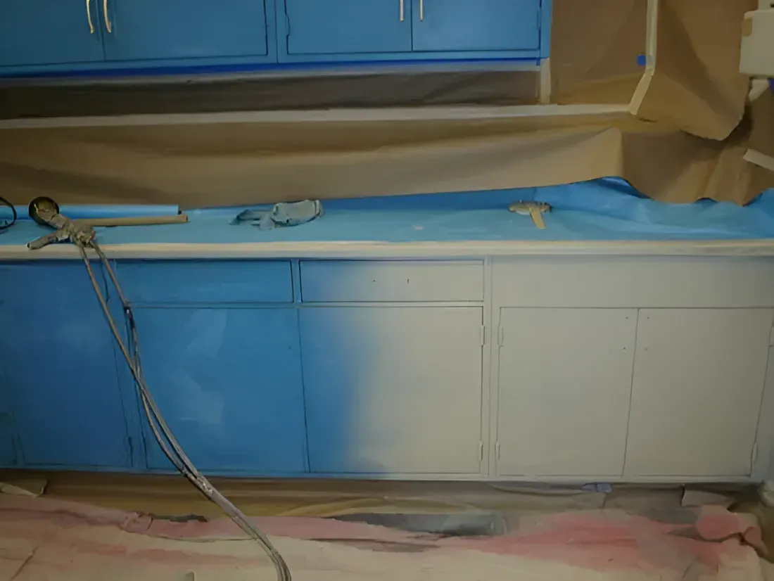 Cabinet painting in progress; lower cabinets half blue, half white. Masking and spray gun visible.