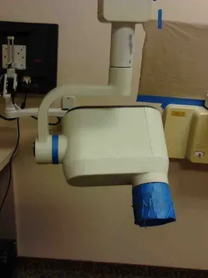 White dental X-ray machine arm and head, blue tape covers, mounted on a wall in a medical setting.