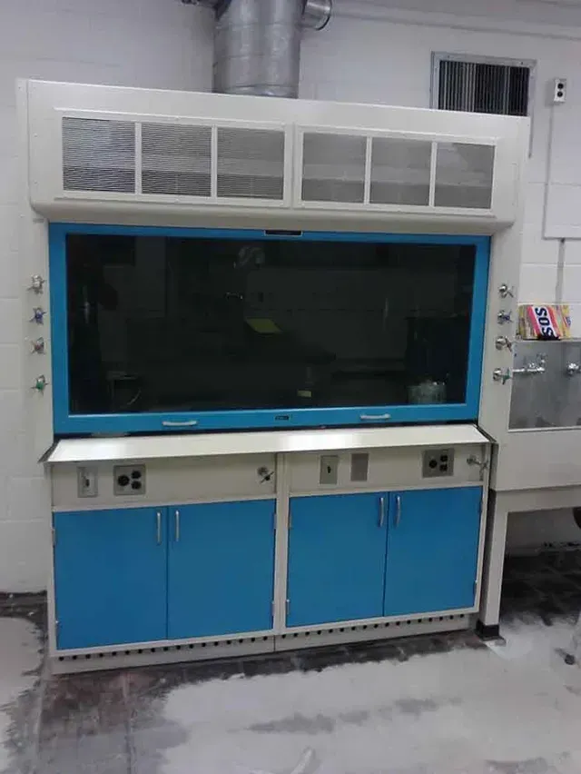 A laboratory fume hood, white with blue accents. The ventilation system is connected to the ceiling.