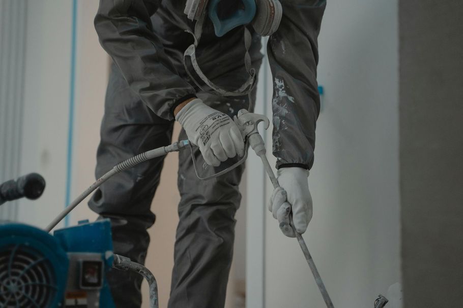 Person in protective suit and gloves using paint sprayer on a wall.