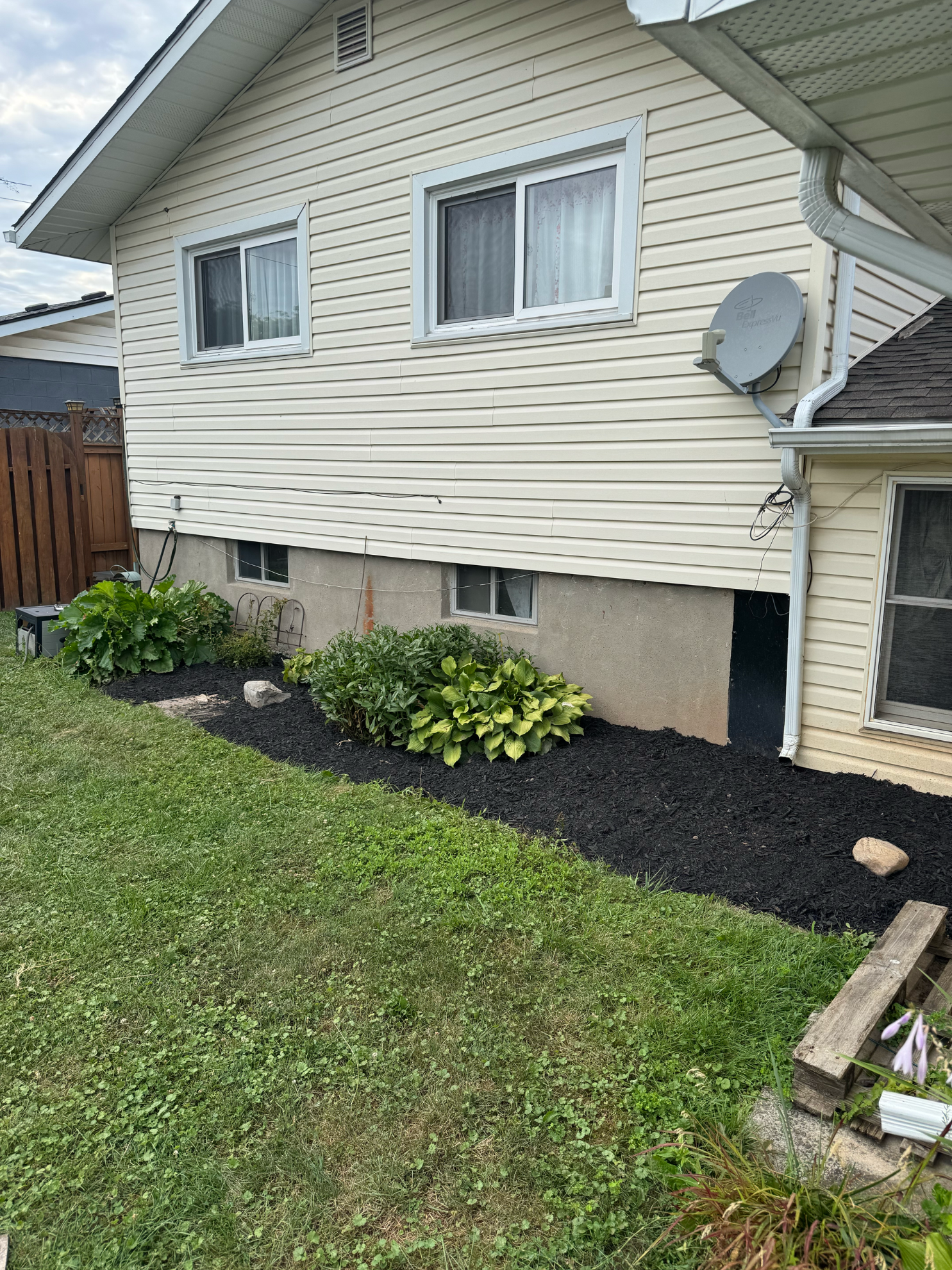 A side view of a cream-colored house with siding, two upper windows, a foundation, a garden bed, and a satellite dish.