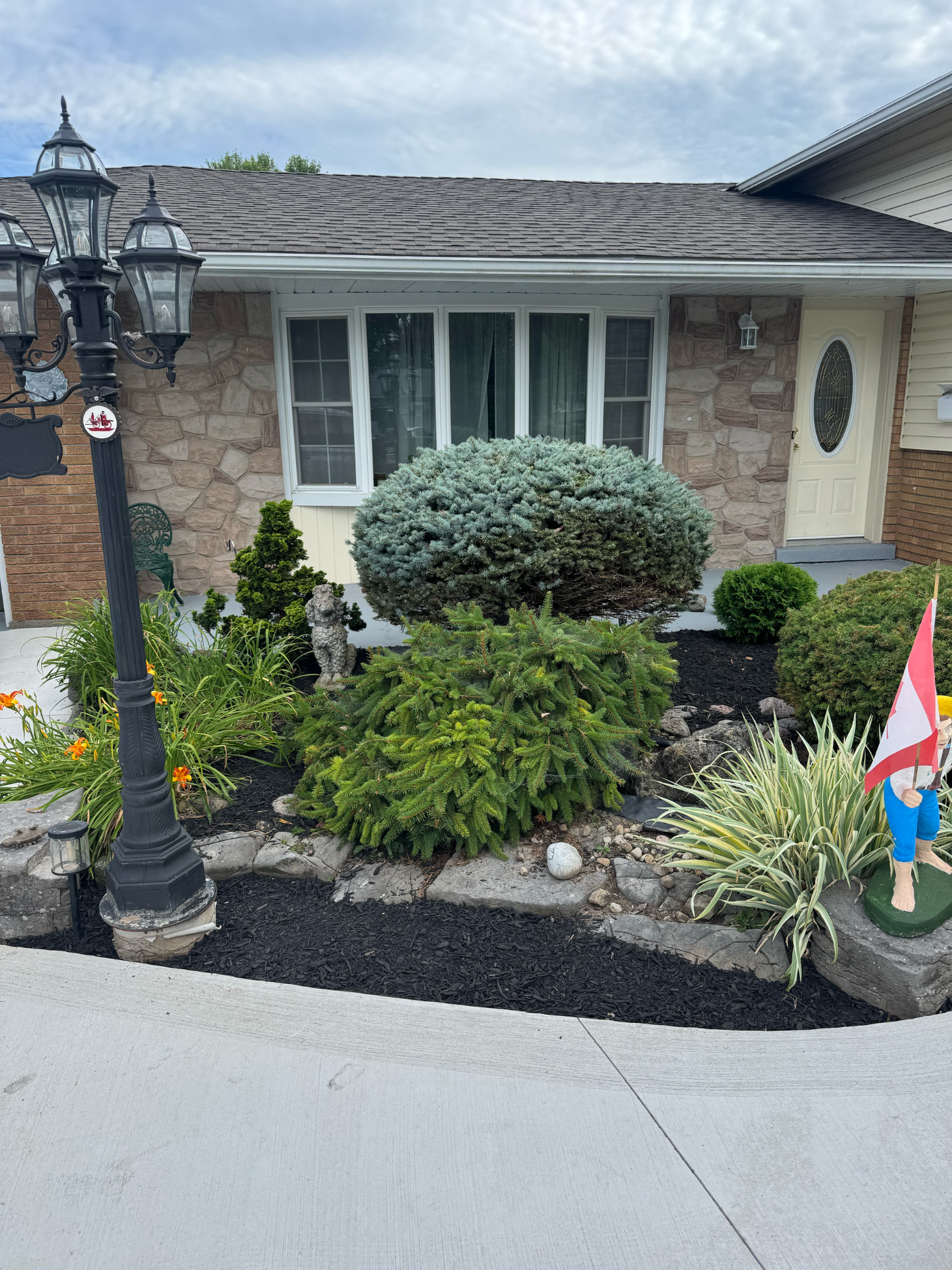 A stone-facade house exterior with a multi-globe street lamp, various green shrubs, and black mulch in the front yard.