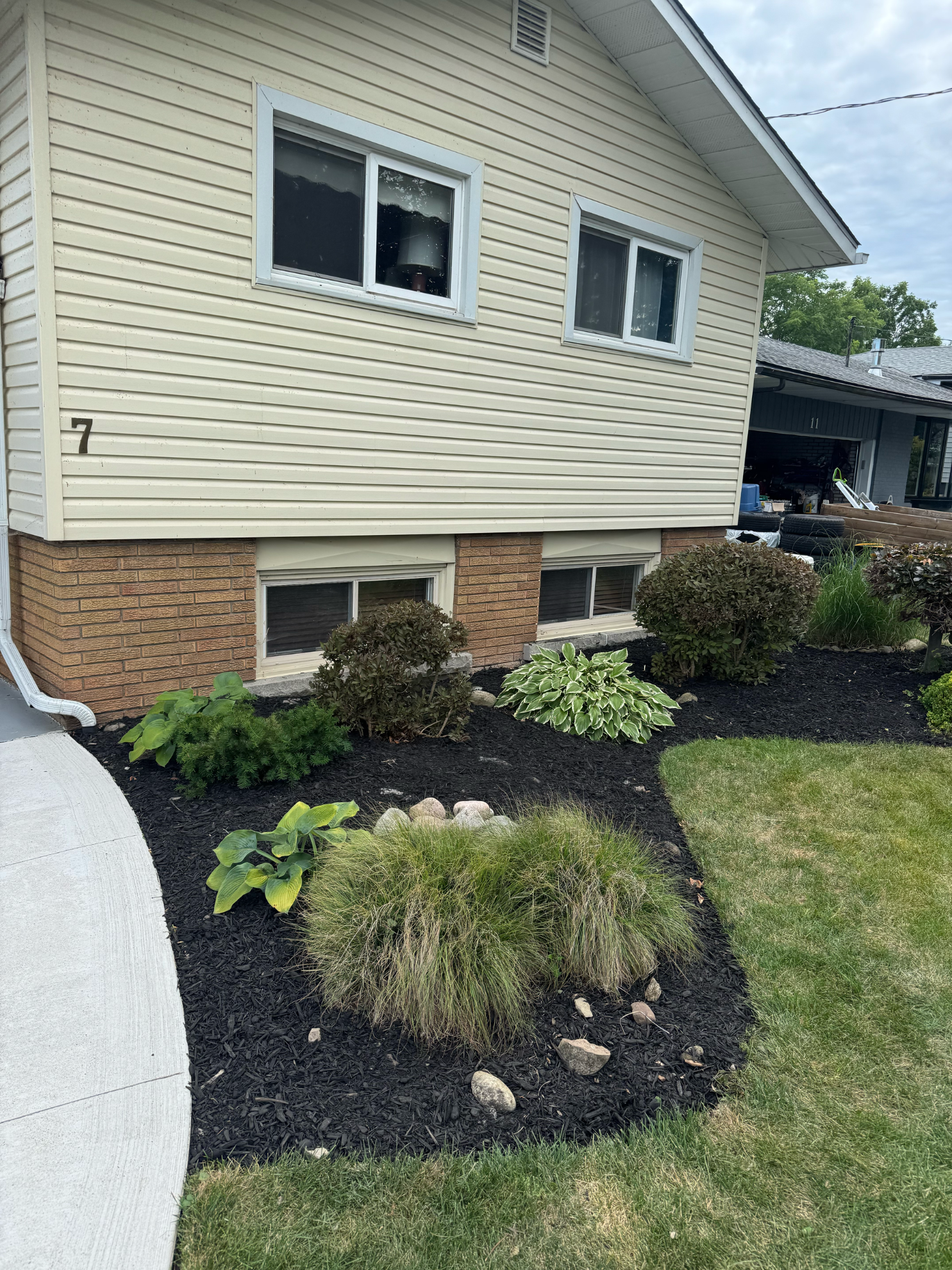 A house with beige siding and a brick foundation, featuring a landscaped garden bed with mulch and assorted green plants.