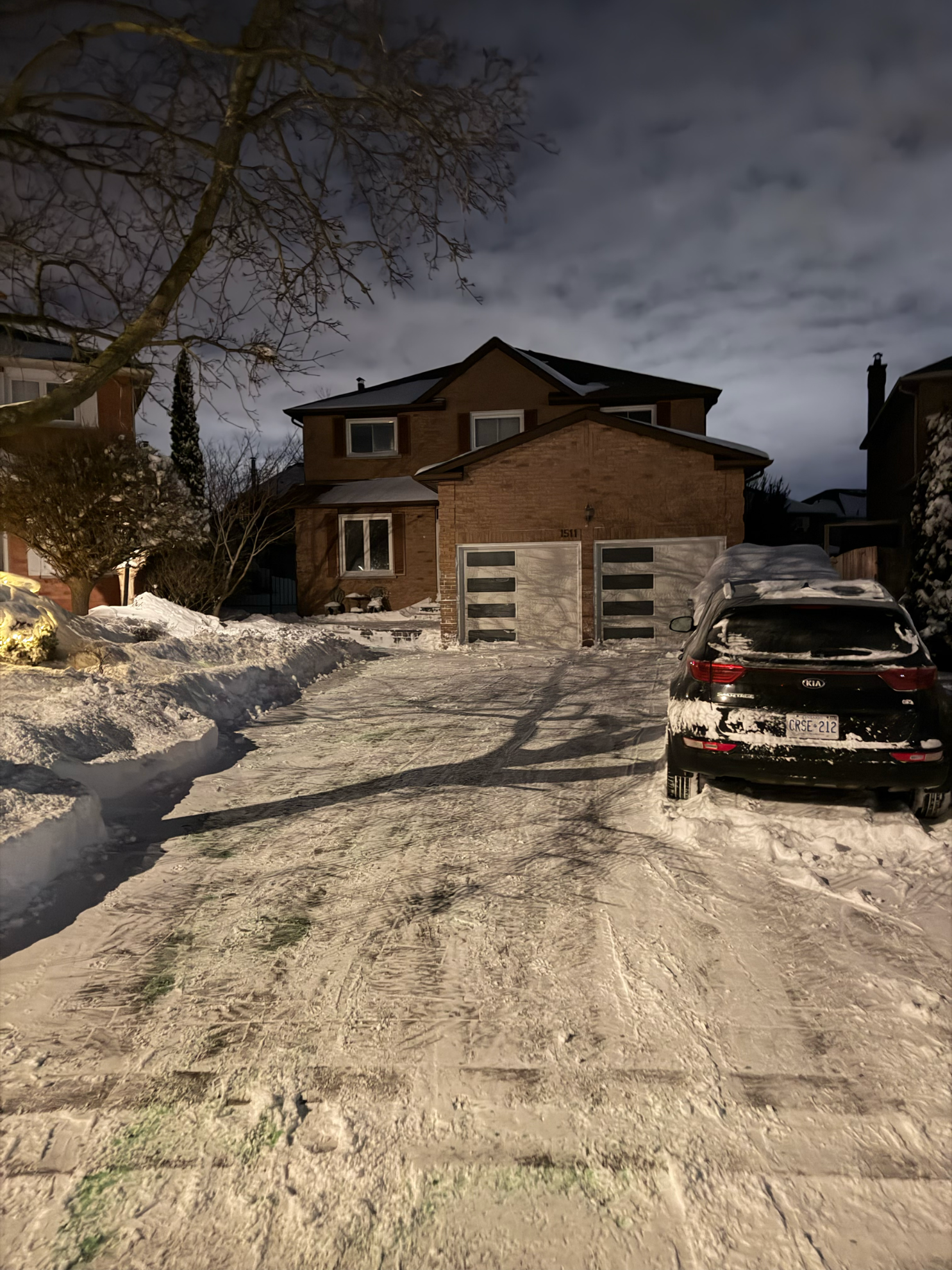 A snowy driveway in front of a brick house at twilight, with a dark SUV parked on the right.