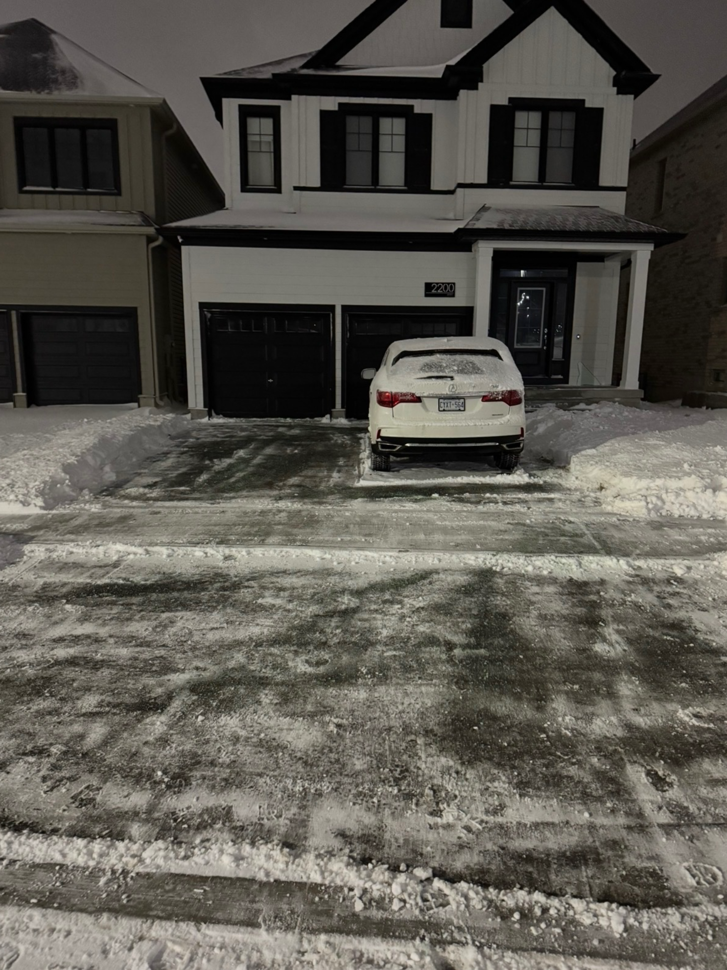 A white two-story house with black trim and a double garage, with a snow-covered driveway and a parked white car at night.