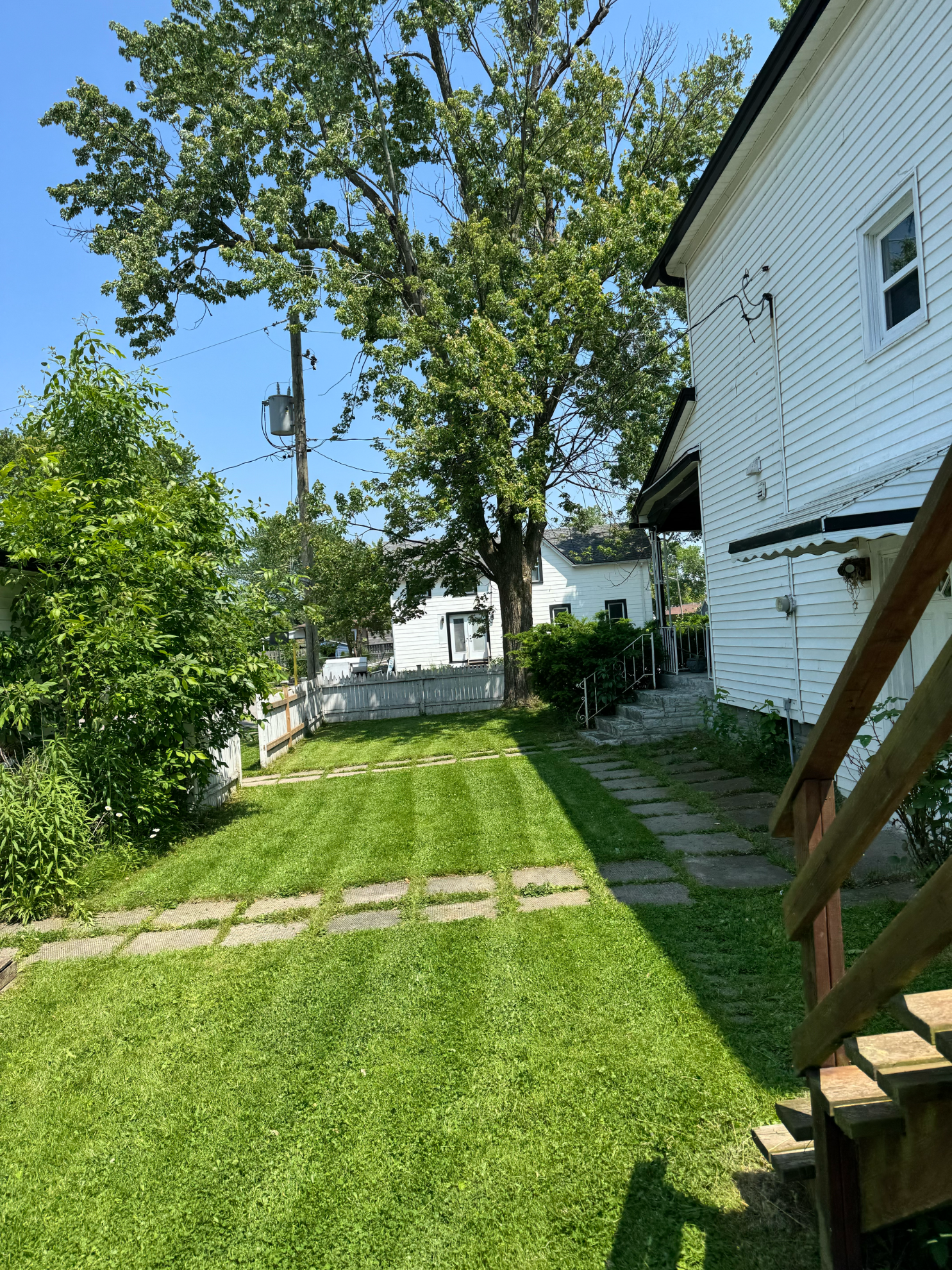 A sunny backyard with green grass, stone pavers, a white house on the right, and a tree against a clear blue sky.
