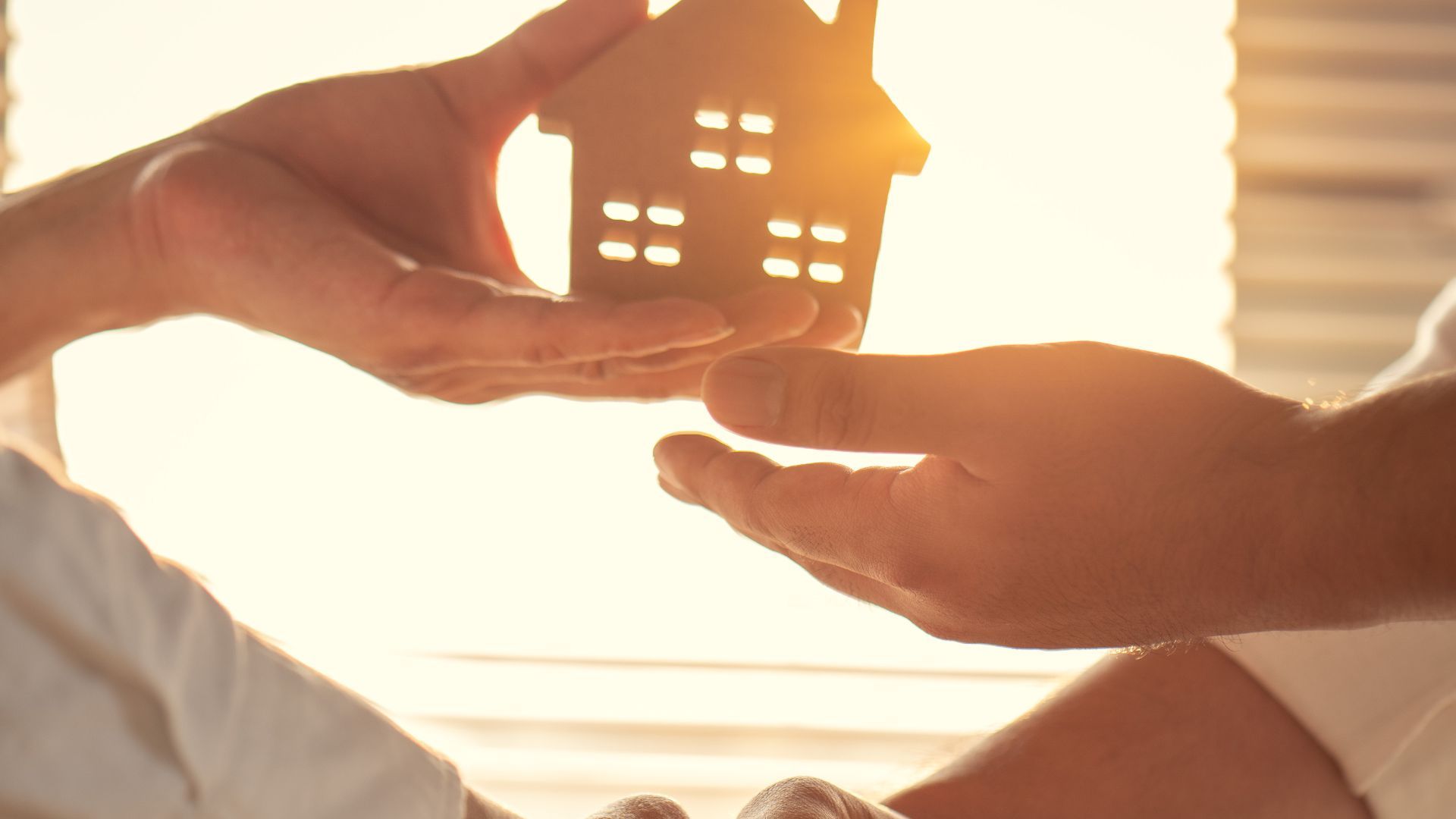Hands exchanging a small wooden house figurine, backlit by sunlight.