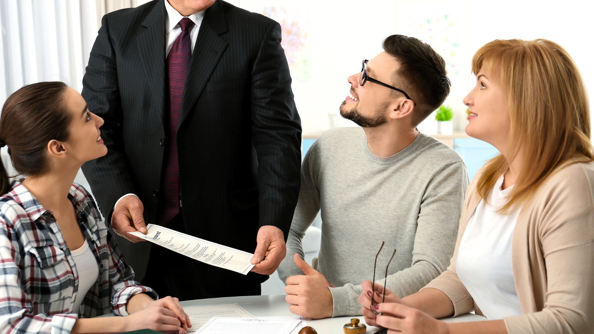 Four people at a table reviewing documents. A man in a suit holds papers; others look on with interested expressions.