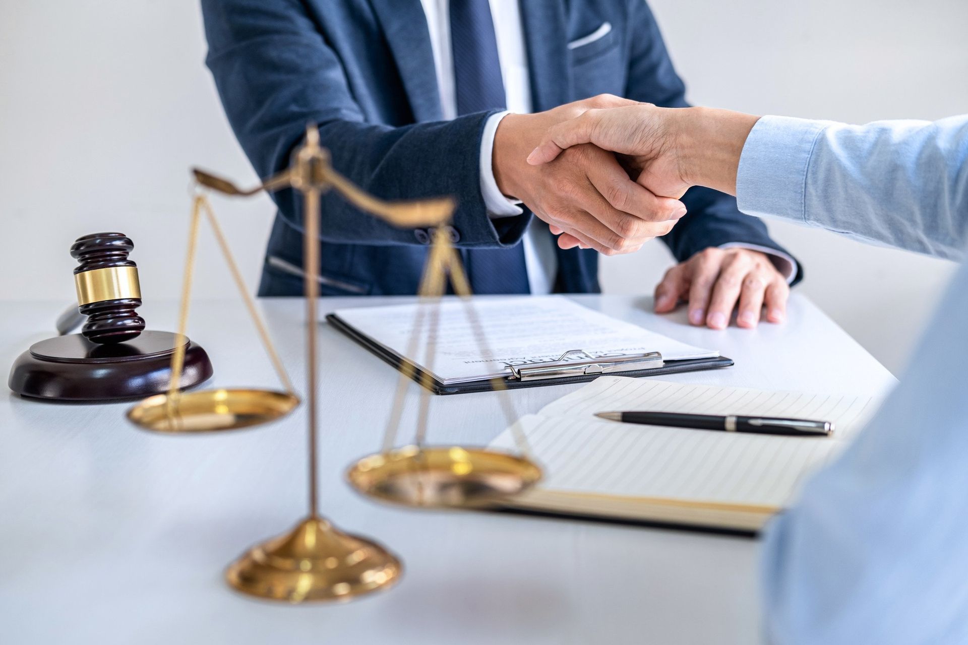 Two people shaking hands near scales of justice, gavel, and documents on a desk.