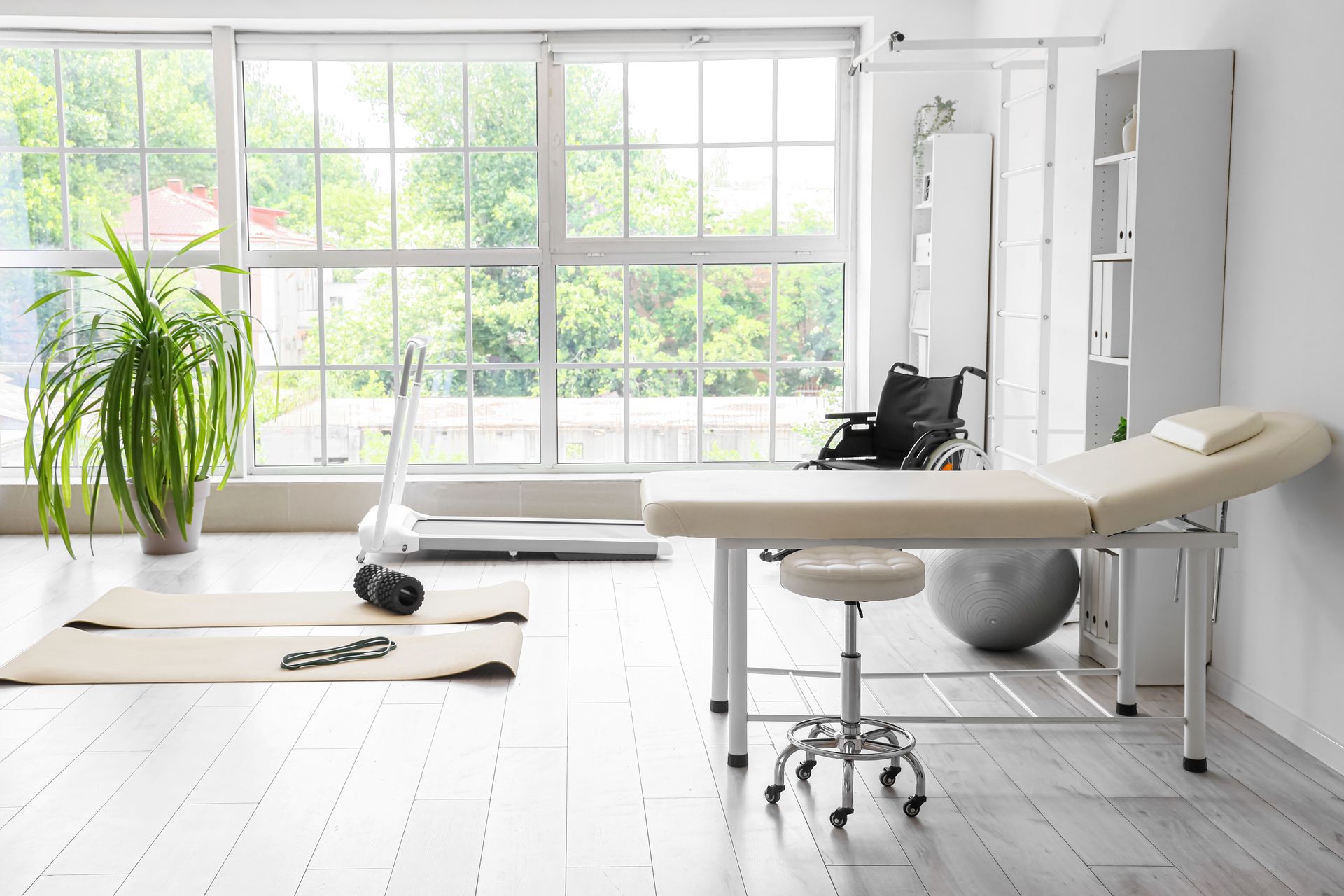 A bright physiotherapy or medical clinic room with a treatment table, exercise mats, a treadmill, and a wheelchair. A bright physiotherapy or medical clinic room with a treatment table, exercise mats, a treadmill, and a wheelchair.
