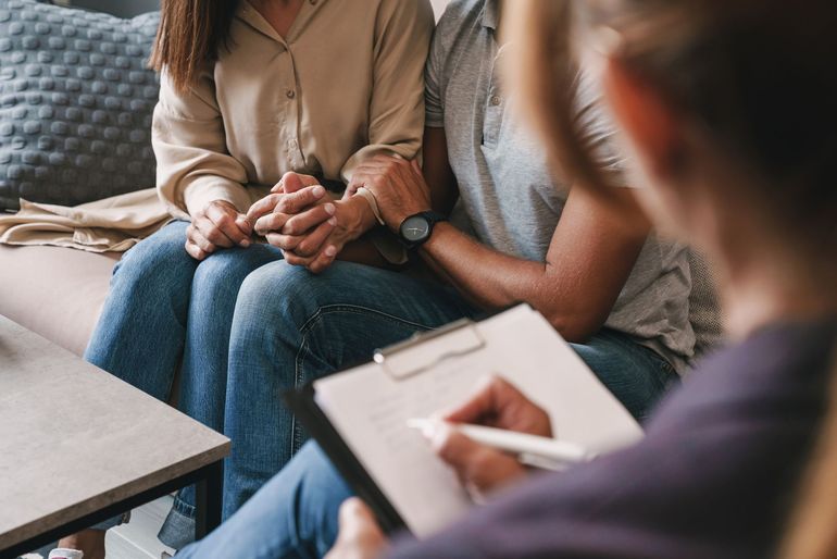 A therapist holds a clipboard while a couple sits together on a couch, holding hands during a counseling session.