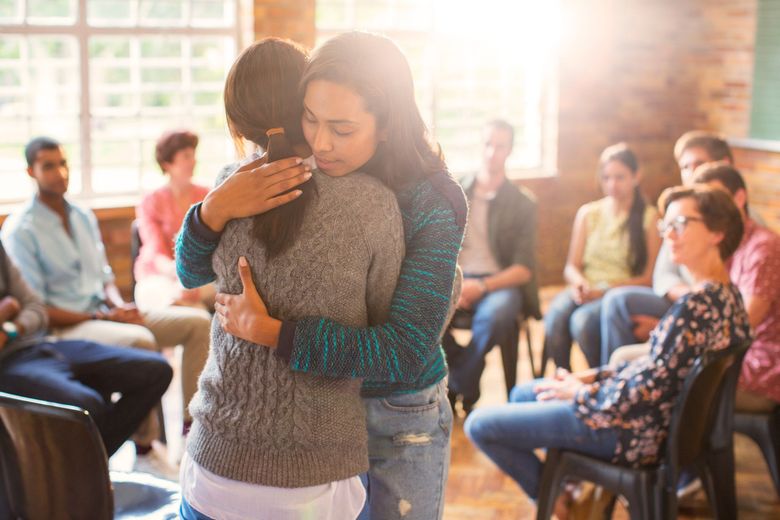 Two people embrace in a group support setting, with others seated in the background in a brightly lit room.