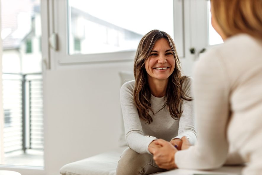 Two people sitting in a bright room, smiling and holding hands in a supportive, comforting gesture.