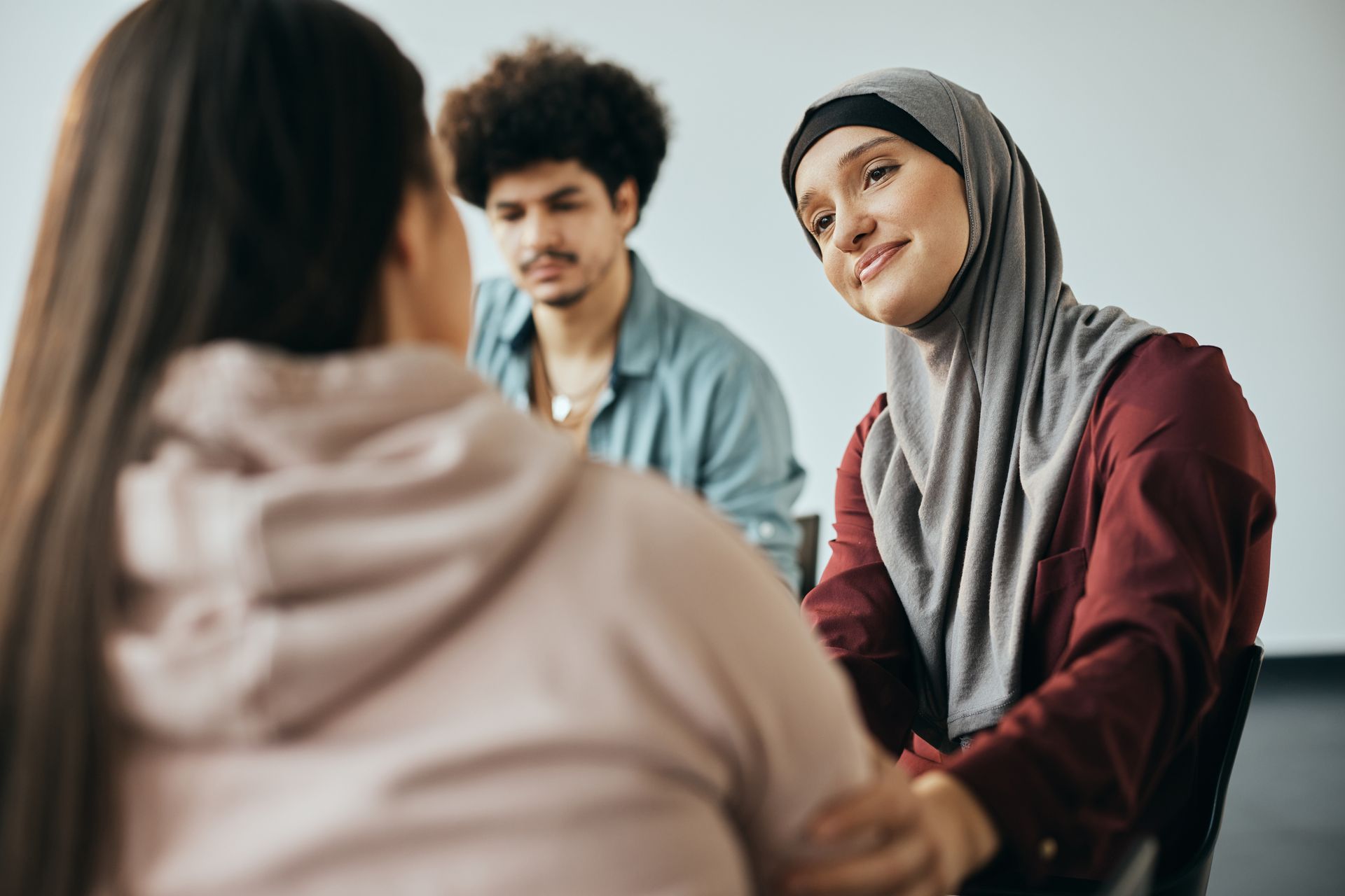 Three people seated in a group, with one person in a hijab smiling while touching the arm of the person in the foreground. Three people seated in a group, with one person in a hijab smiling while touching the arm of the person in the foreground.