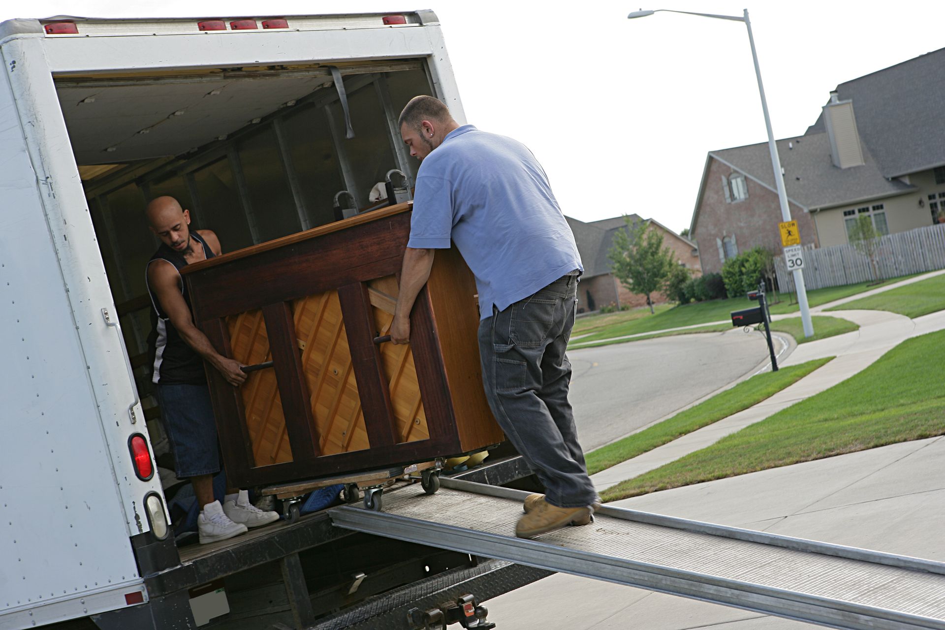 Two people loading a piano into a truck using a ramp on a quiet residential street.
