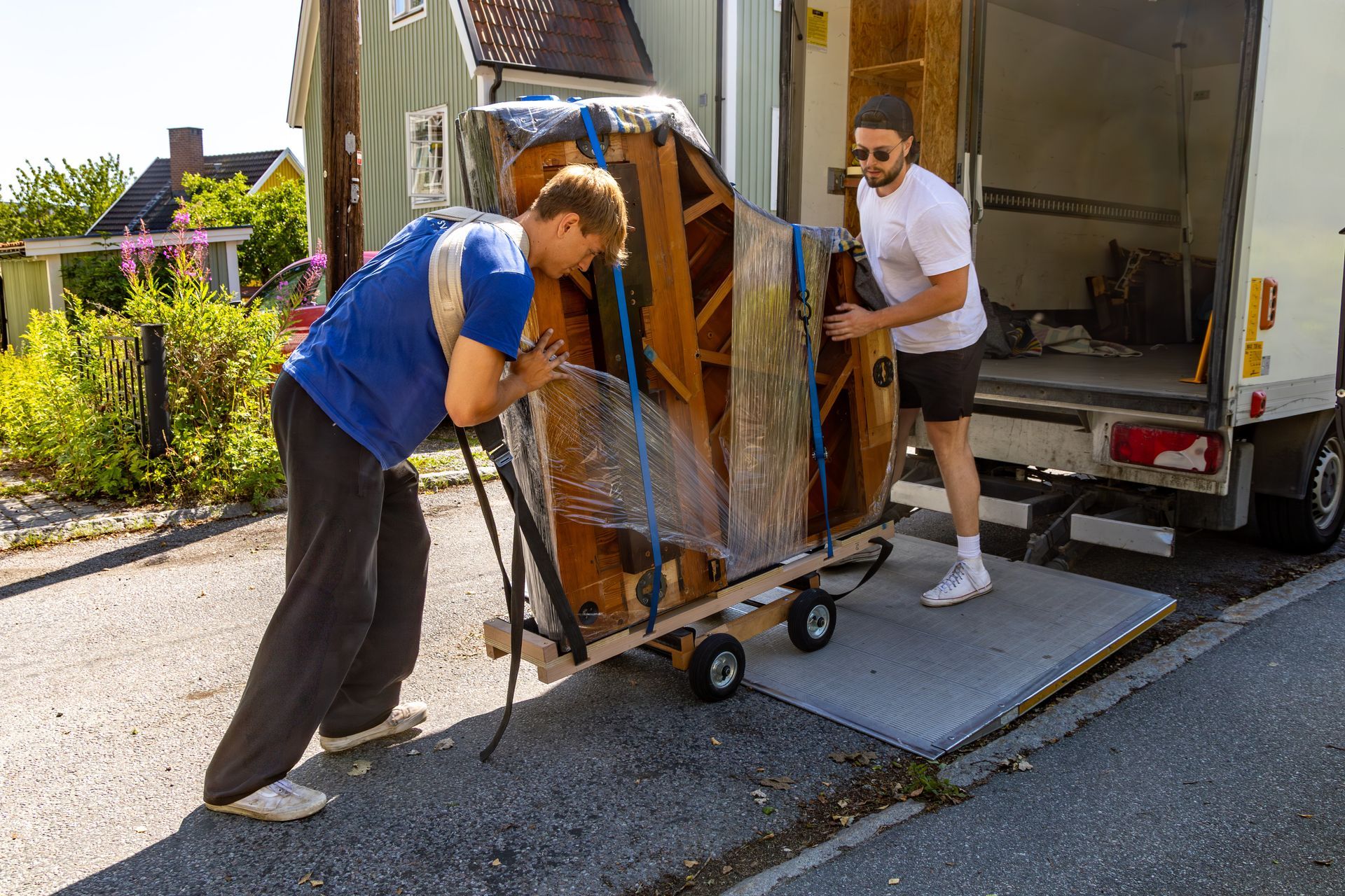 Piano movers carefully loading upright piano onto dolly and truck ramp outside home.