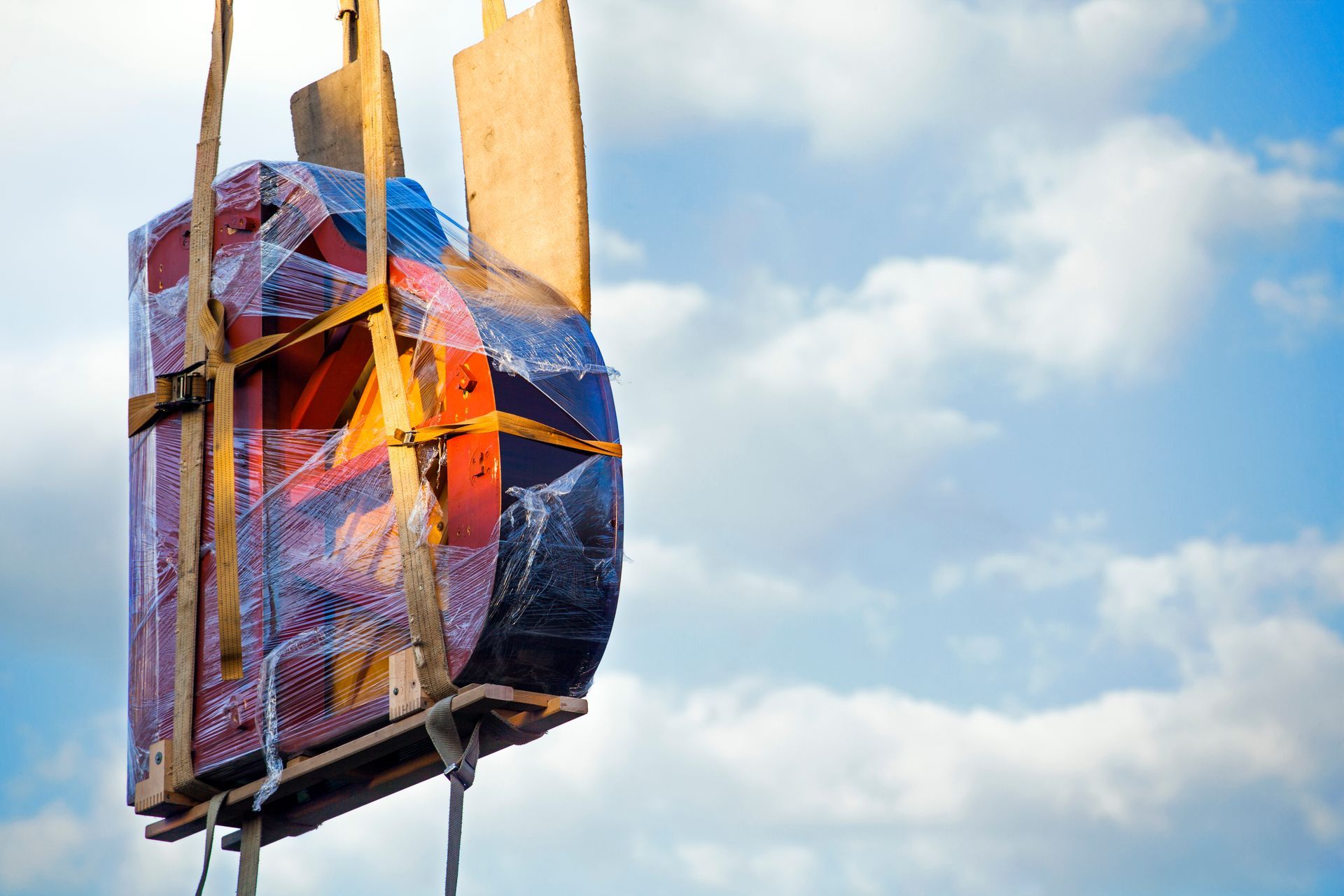 Heavy industrial equipment strapped to a pallet and wrapped in plastic, lifted by a crane against a blue sky. Heavy industrial equipment strapped to a pallet and wrapped in plastic, lifted by a crane against a blue sky.