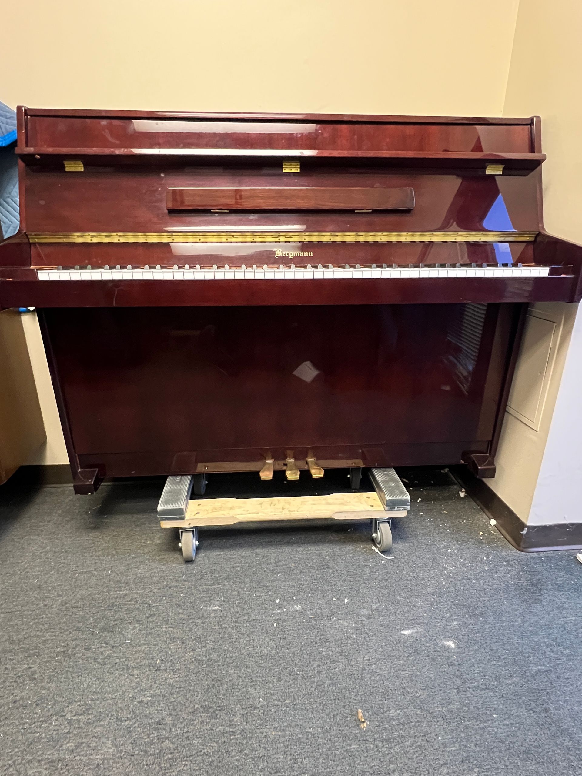A maroon upright piano on a rolling platform in a room with a beige wall and dark carpet.