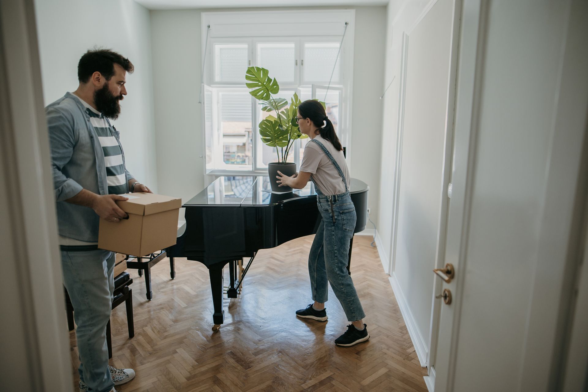 Woman places plant on piano as couple unpacks boxes in their sunlit new apartment.