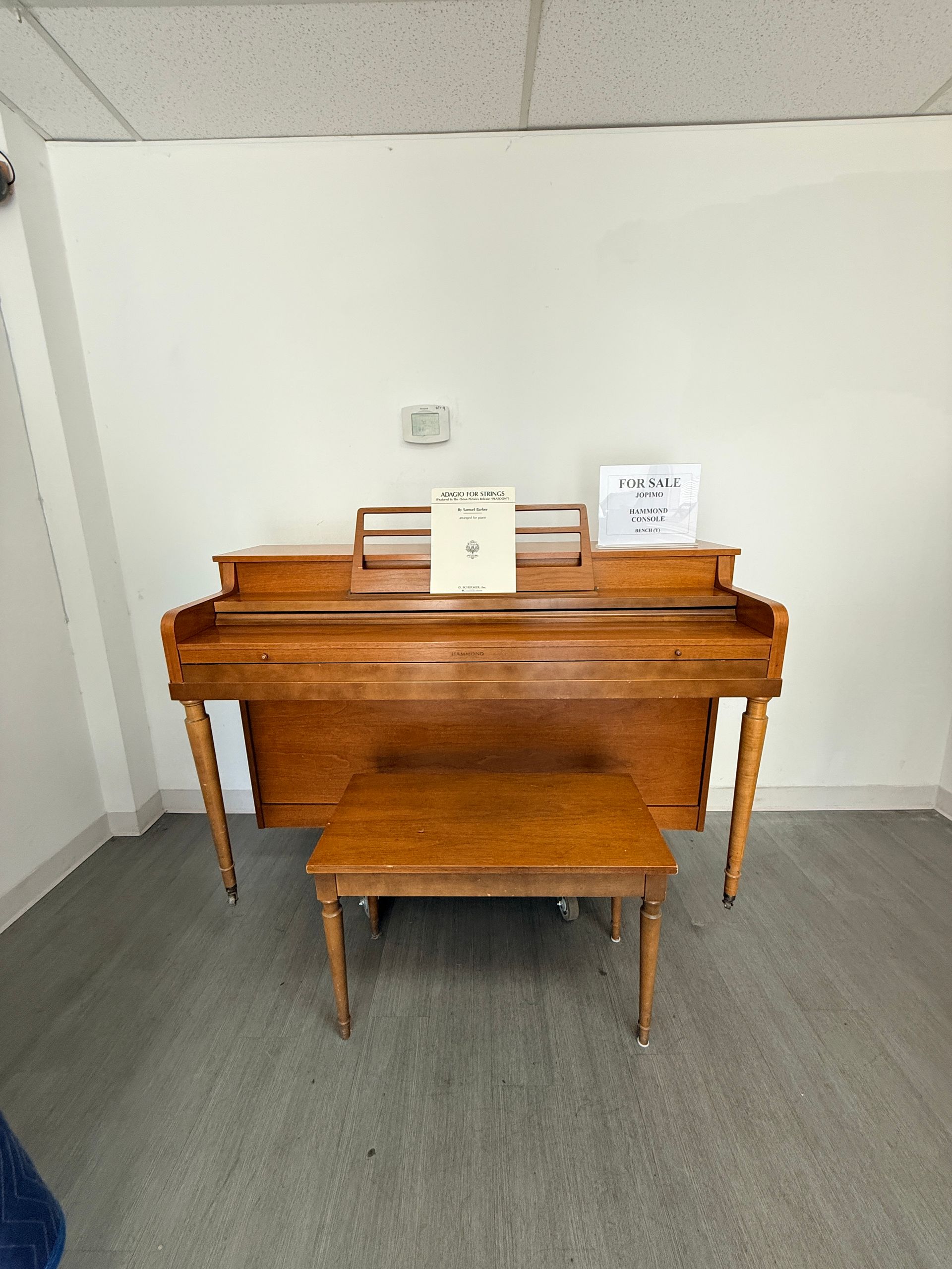 Wooden upright piano with bench in a plain hallway, with small signs on top.