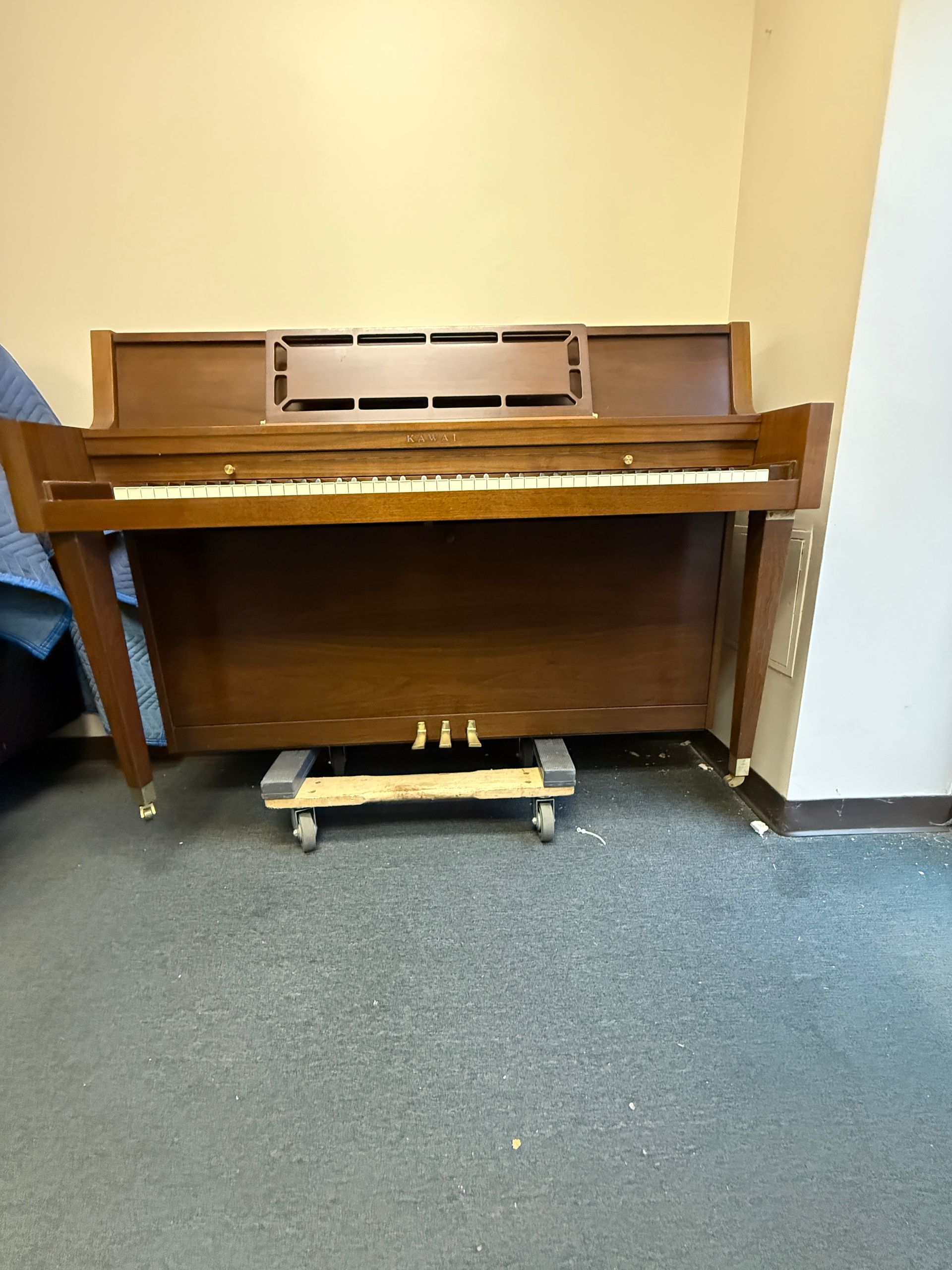 Wooden upright piano on a wheeled dolly against a beige wall and blue carpet.