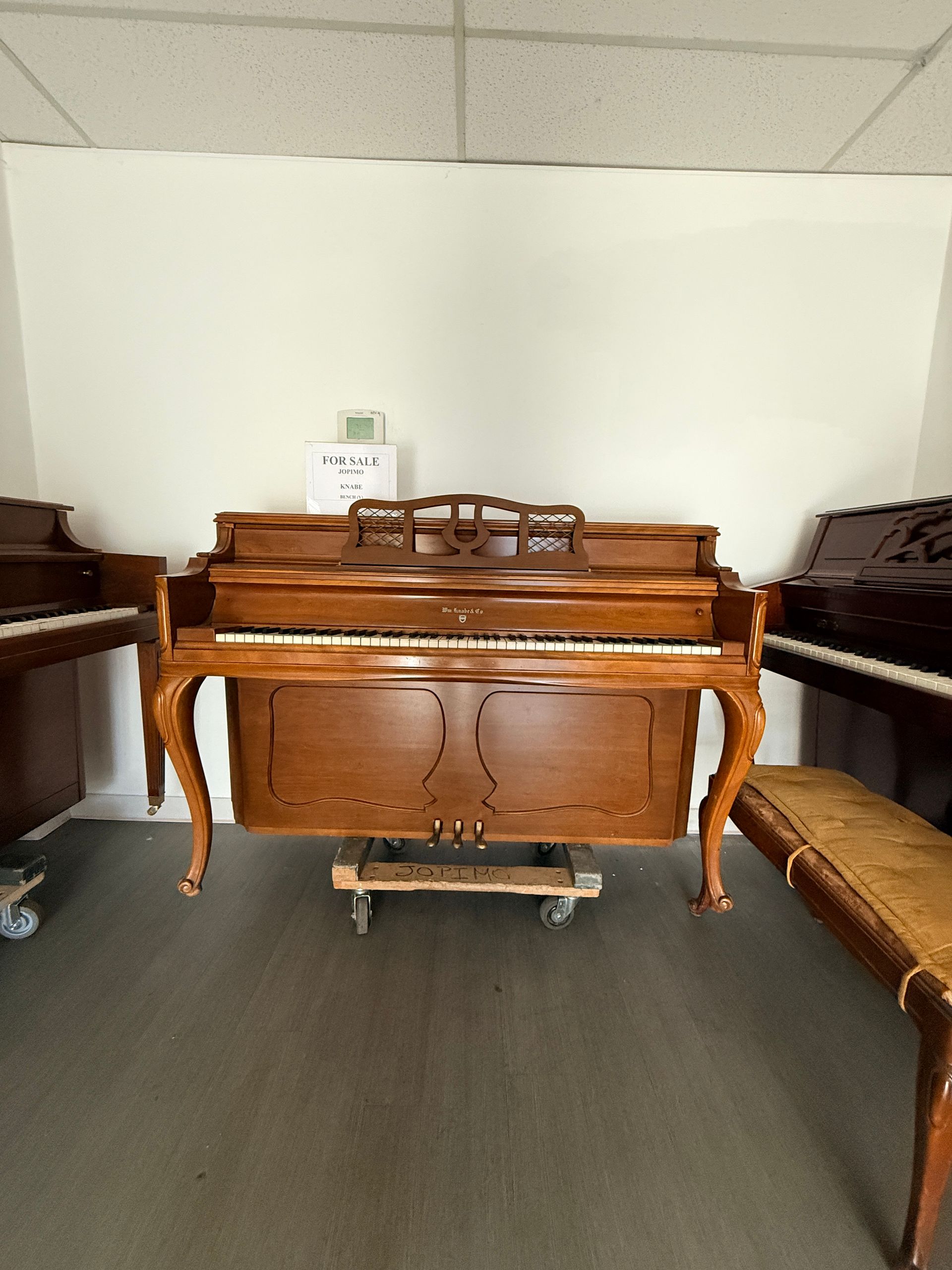 Wooden upright piano on a wheeled dolly in a room with benches and carpeted floor