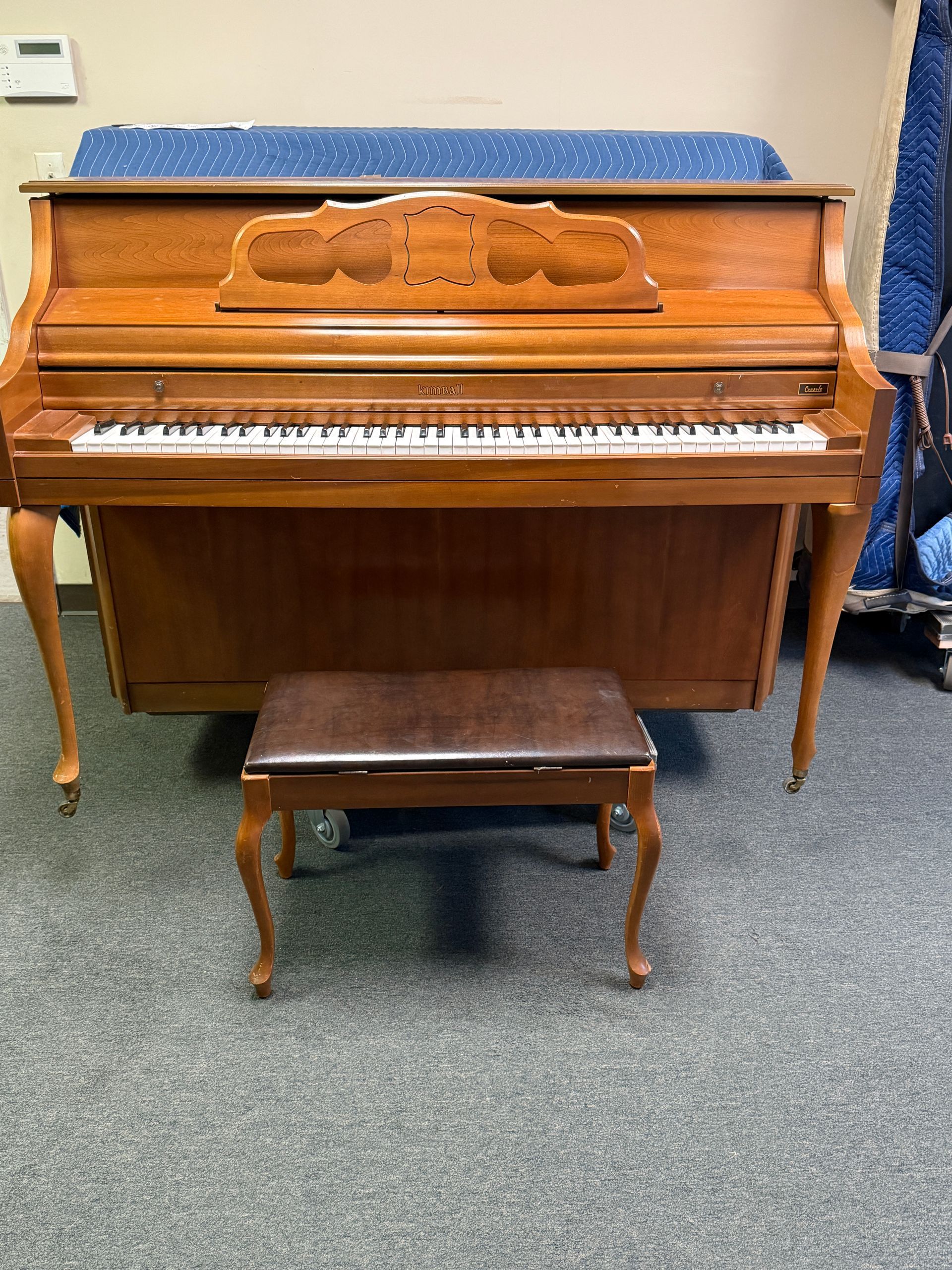 A light brown upright piano with a matching bench on a gray carpet.