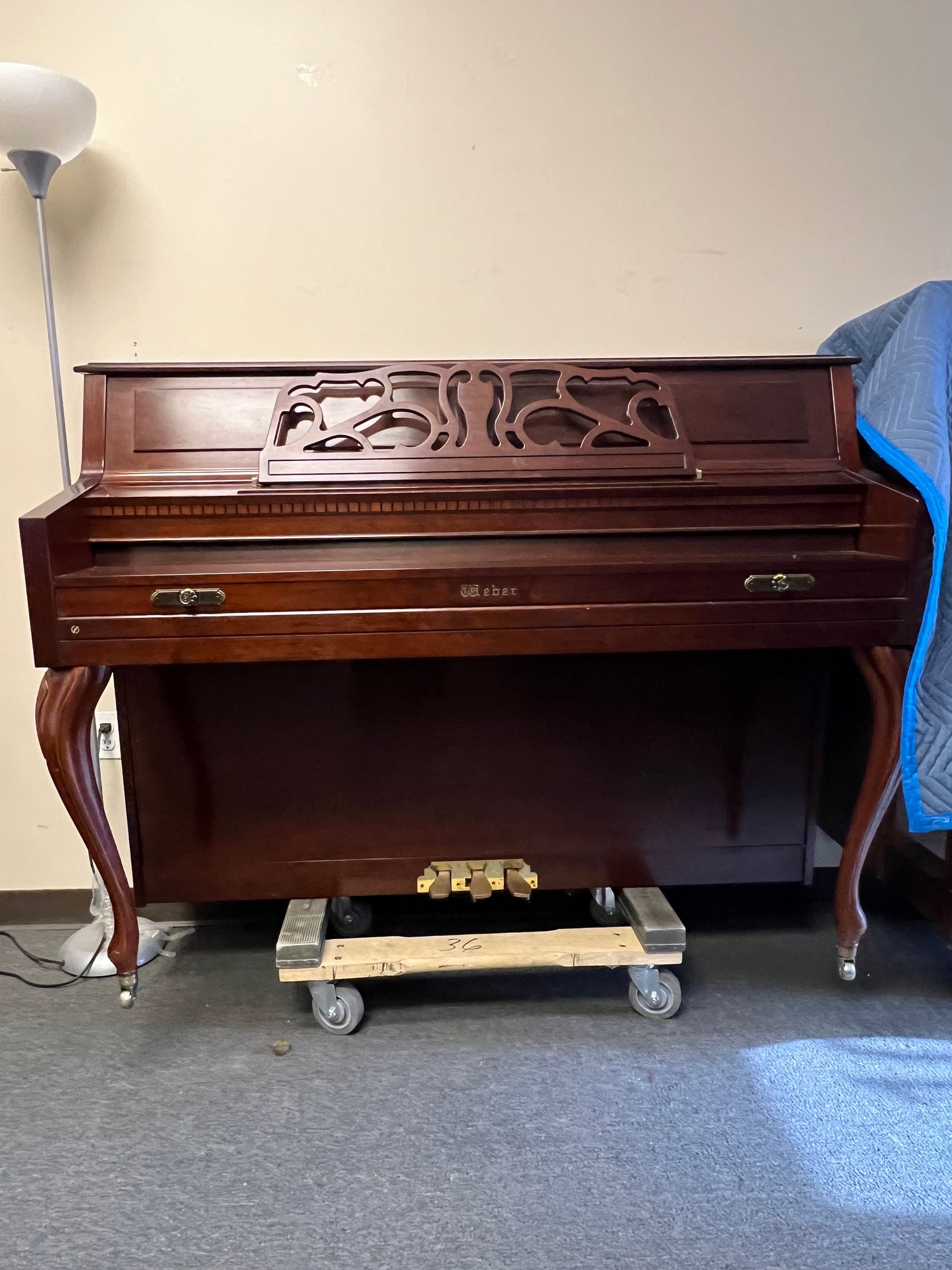 Dark wooden upright piano on a dolly, in a room with a floor lamp and a moving blanket.