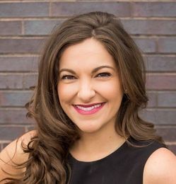 A woman in a black dress is smiling in front of a brick wall.