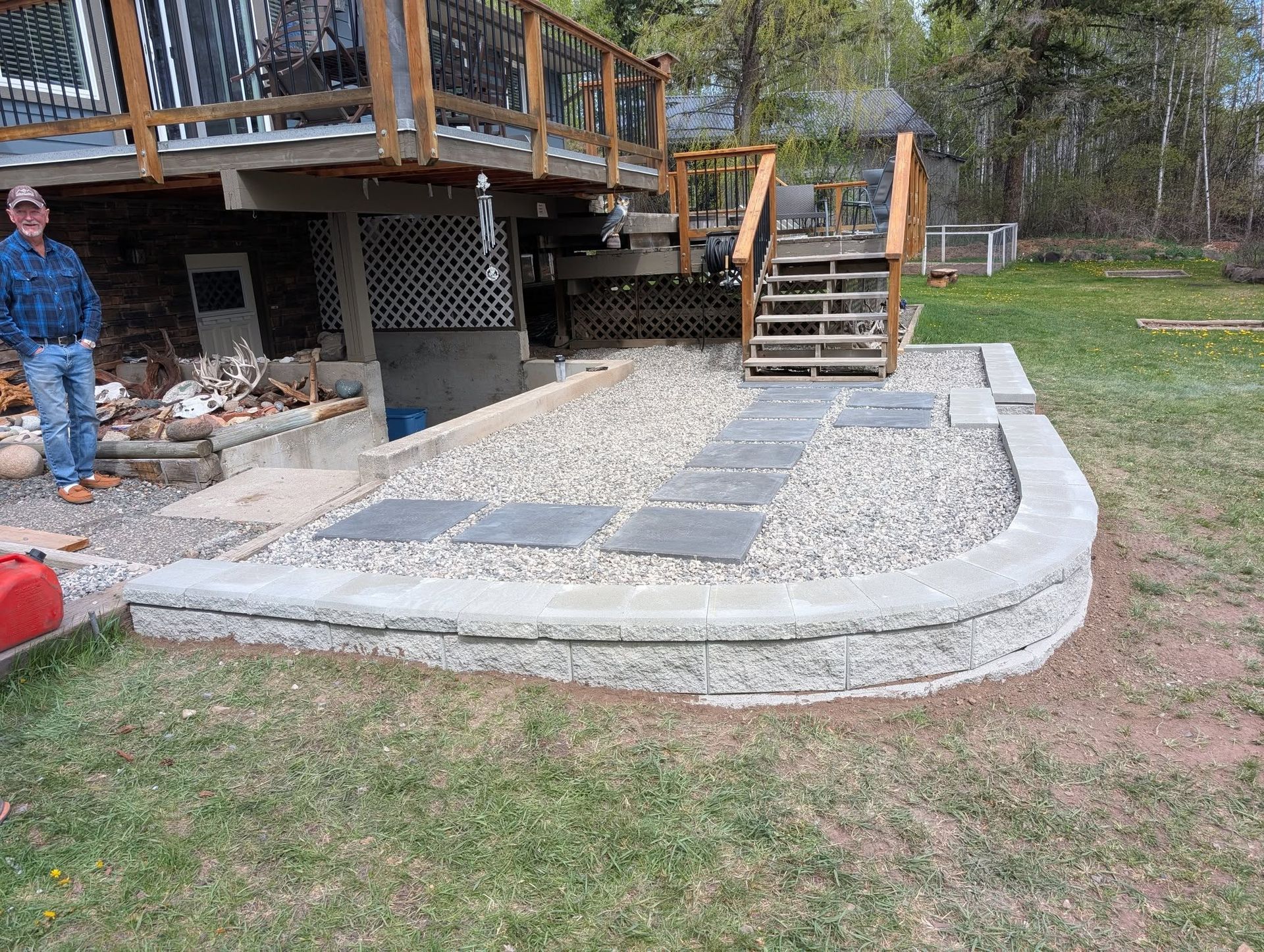 Man stands beside a stone retaining wall, a gravel area, and a deck with stairs.