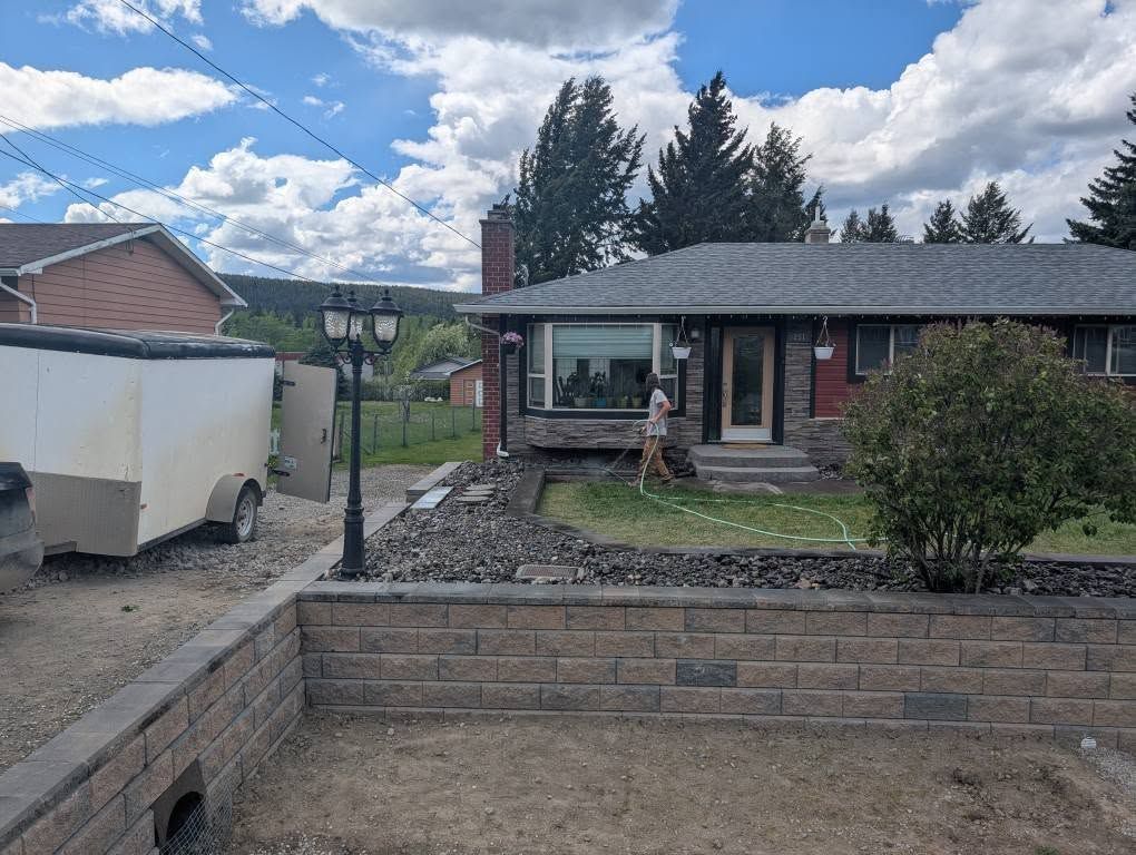A person waters a front yard with a house. A brick retaining wall and trailer are in view.
