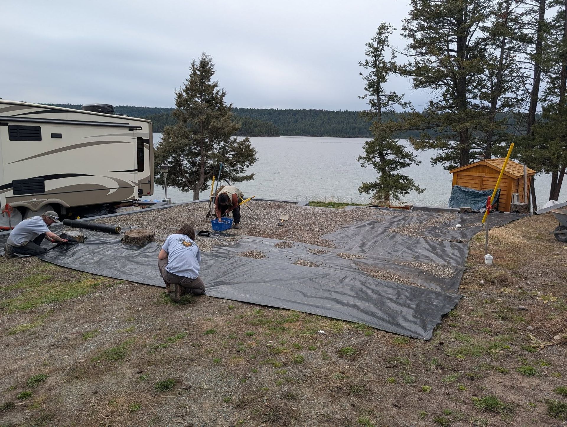 Three people working on a campsite setup near a lake, laying down a ground cover near a RV and a small tent.