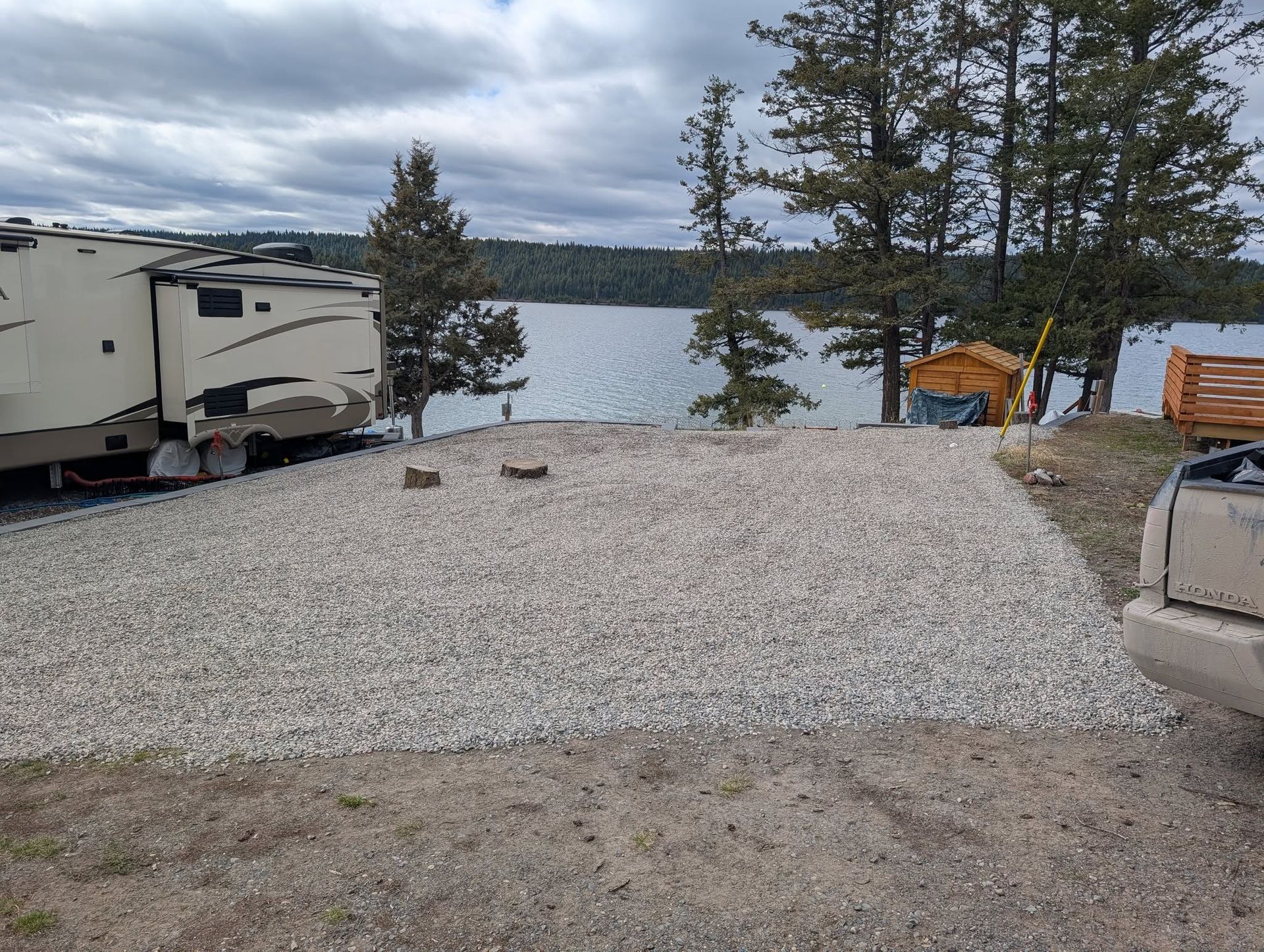 Gravel RV campsite with a lake view, with an RV and trees in the background under a cloudy sky.