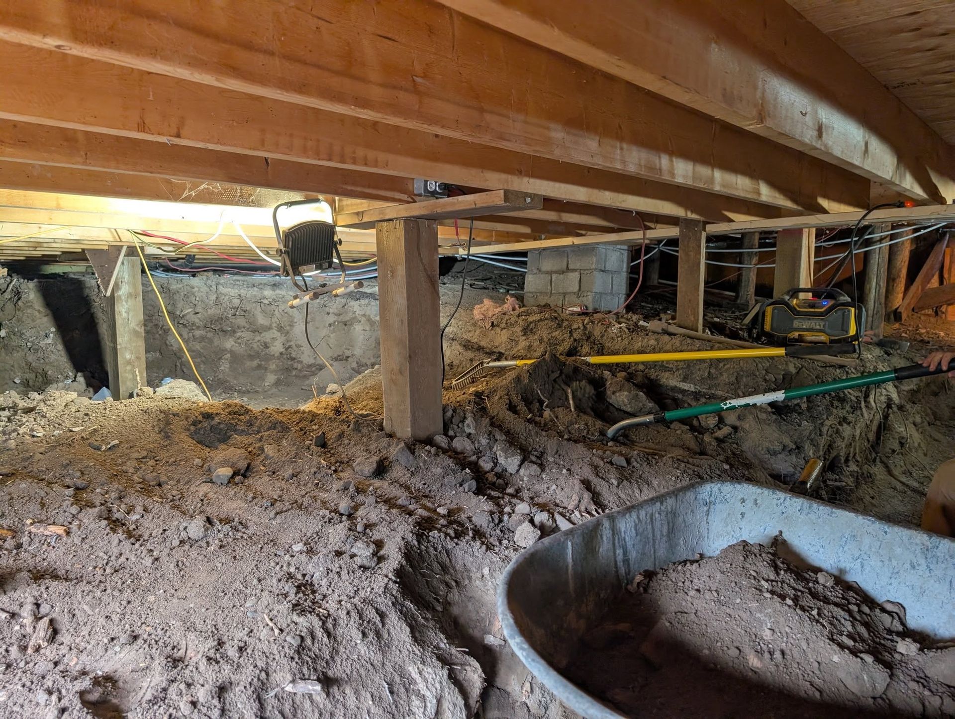 Under-house crawl space with dirt floor, wood beams, support posts, and construction materials;