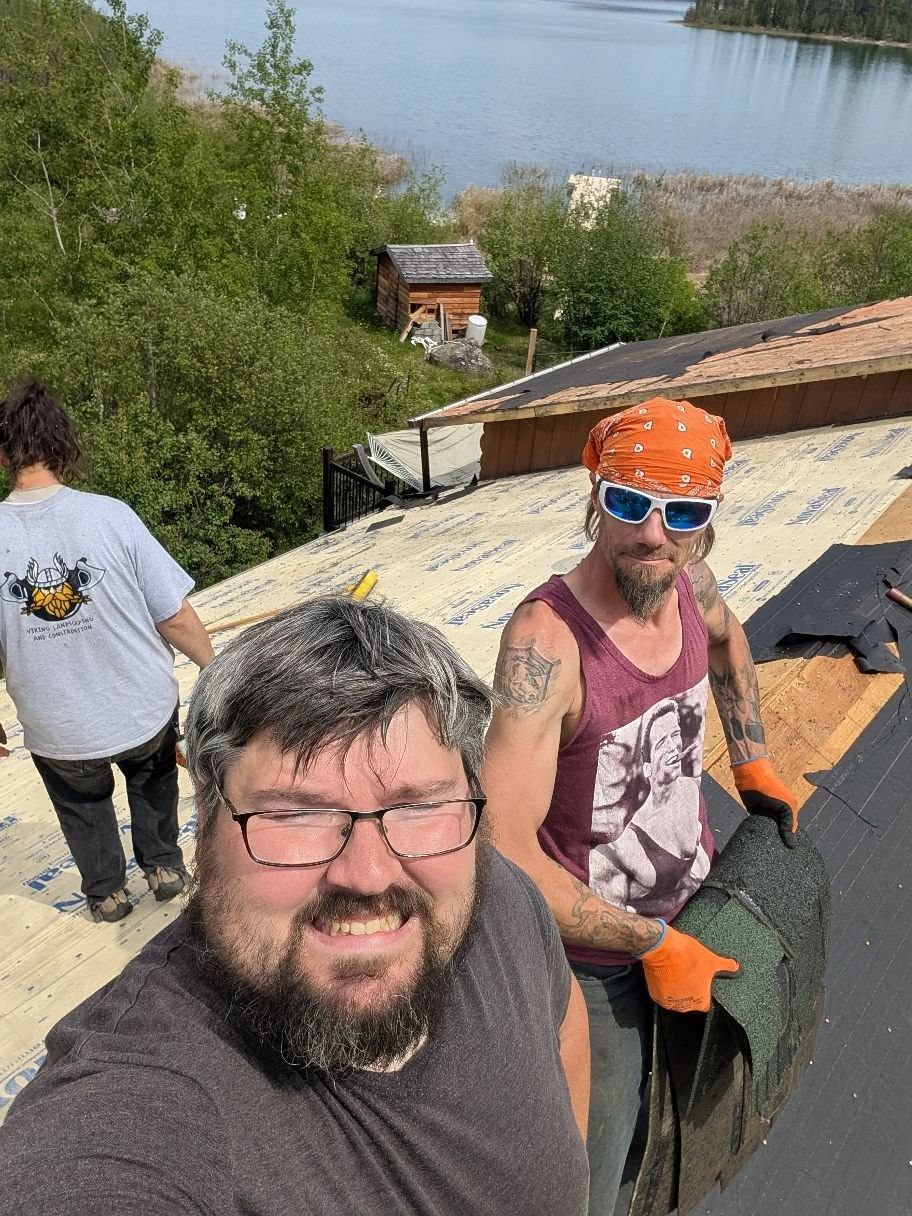 Three men on a roof near a lake. One smiling at the camera, others working with materials. Sunny day.