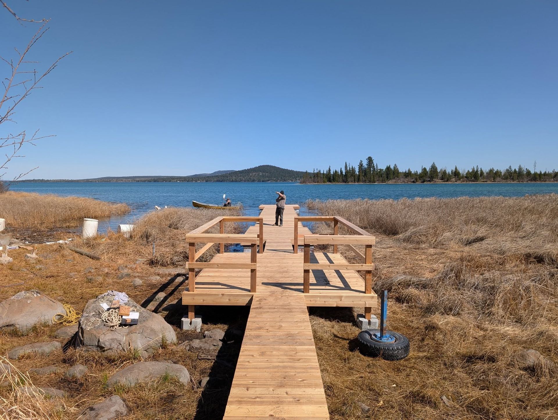 Wooden dock extends into a lake, person stands at the end, clear blue sky above, reeds and rocks in the foreground.