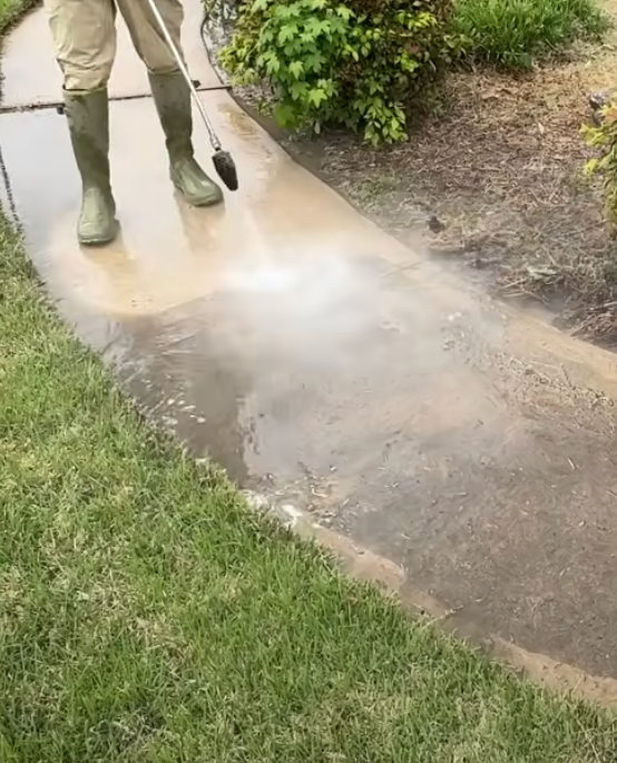Person in boots pressure washing a concrete walkway, near green grass and bushes.