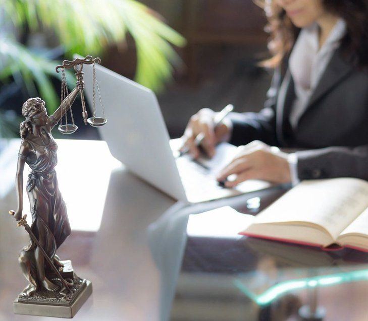 A lady justice statue is sitting on a desk next to a woman using a laptop