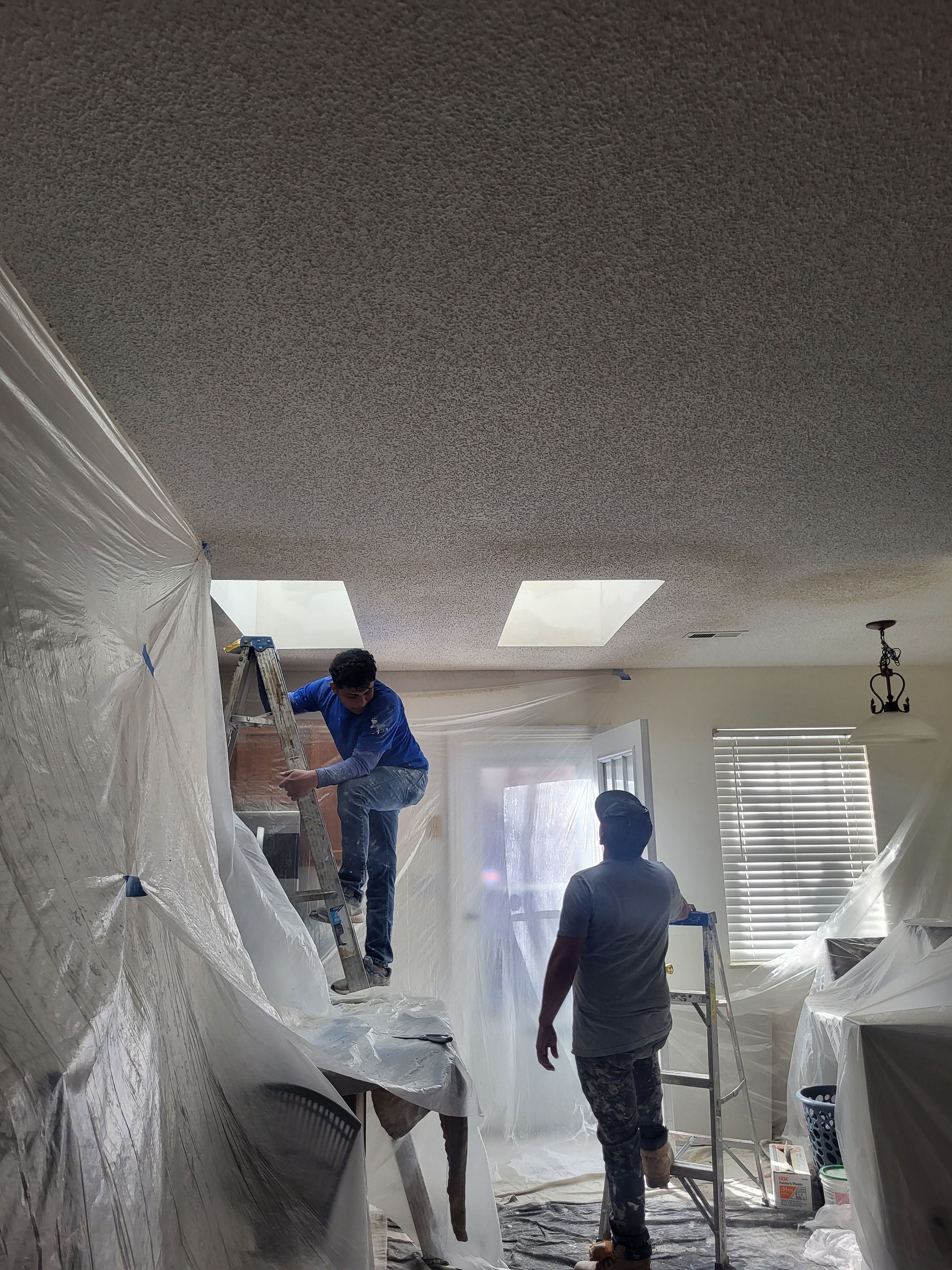 Two men are painting a wall in a room covered in plastic.
