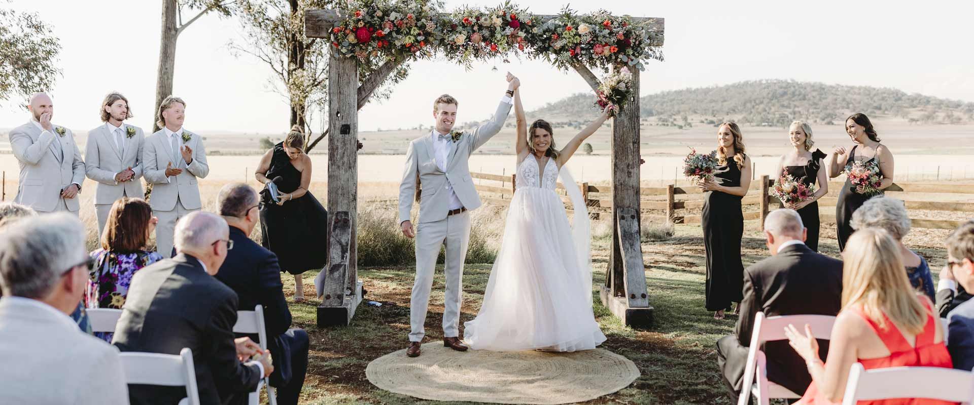 Newlyweds raise arms under a floral arch during an outdoor wedding ceremony. 