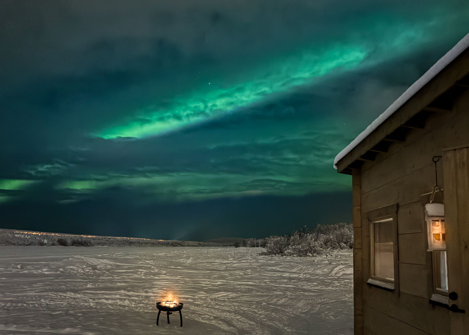 Northern Lights above an outdoor fire next to a wooden cabin in snowy landscape.