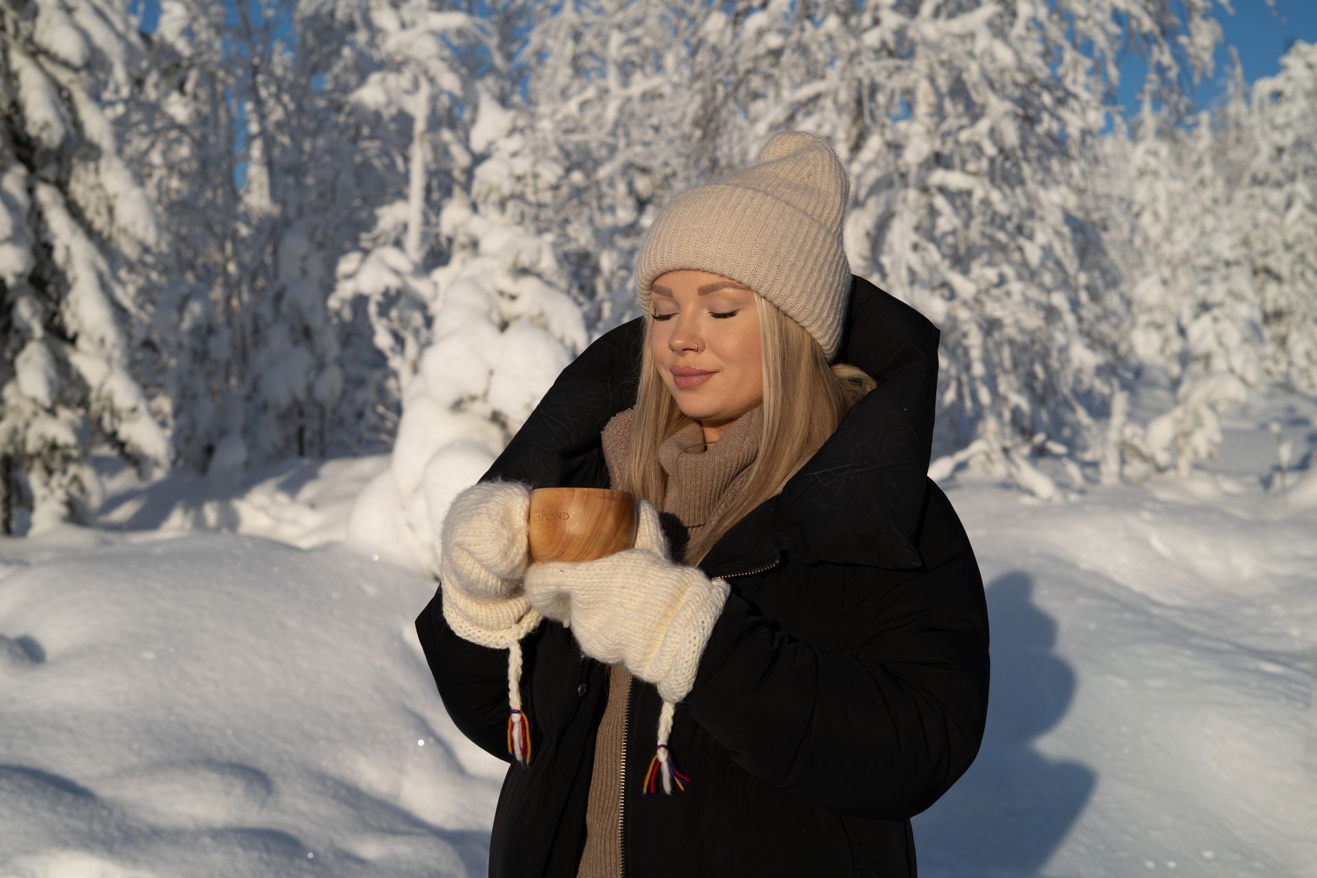 Woman enjoying sunshine while holding a kuksa wooden cup in her hands, surrounded by snow.