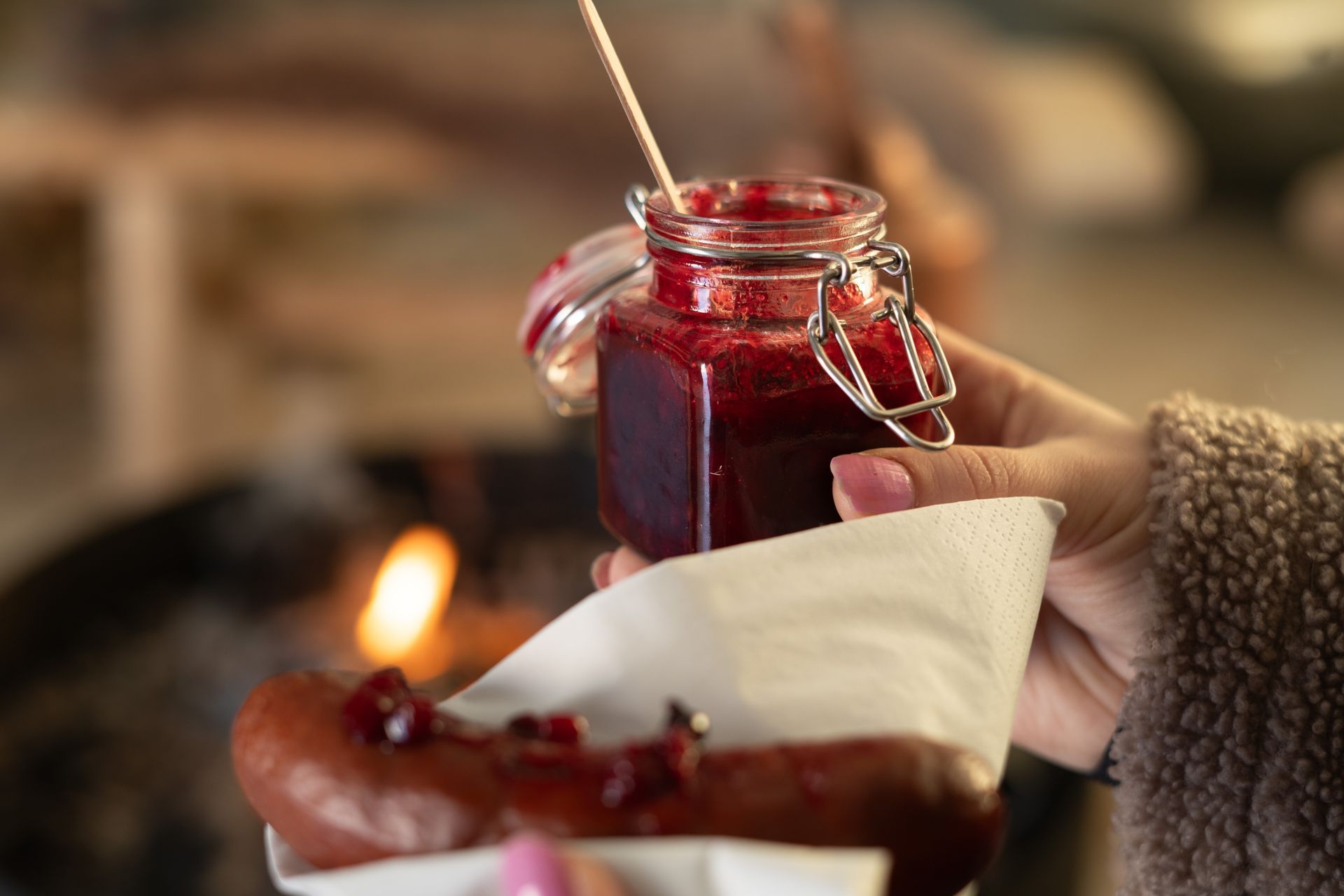 Hand holding a reindeer sausage with lingonberry chutney, in front of a fire.