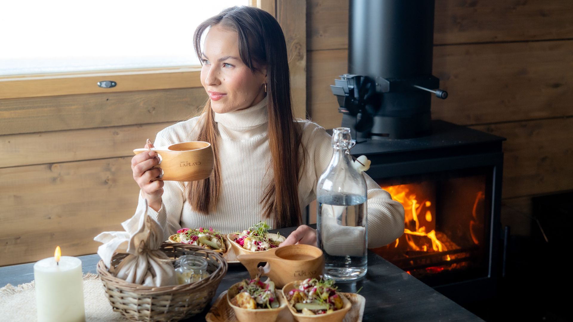 Woman drinking from a kuksa inside a cabin with fire in the background