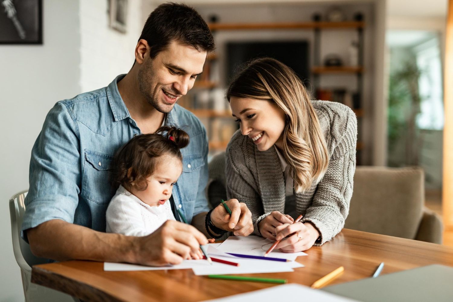 Família de três pessoas colorindo juntas em uma mesa na sala de estar, sorrindo.
