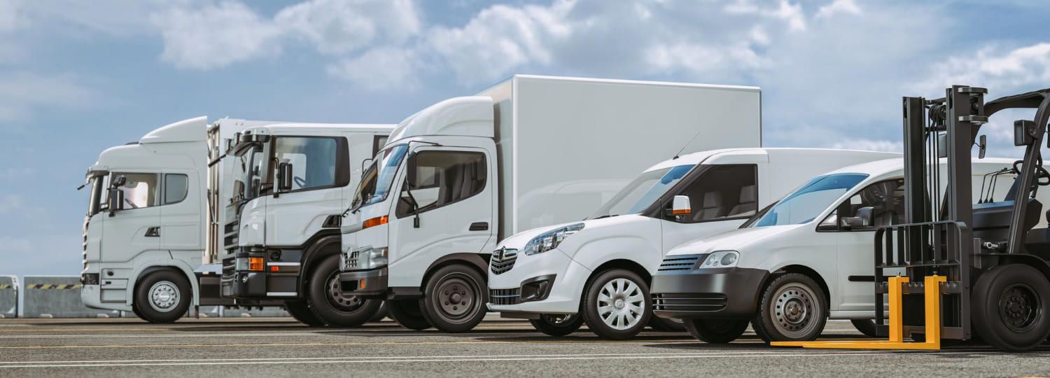 Veículos de entrega brancos alinhados sob um céu nublado, incluindo caminhões, vans e uma empilhadeira.