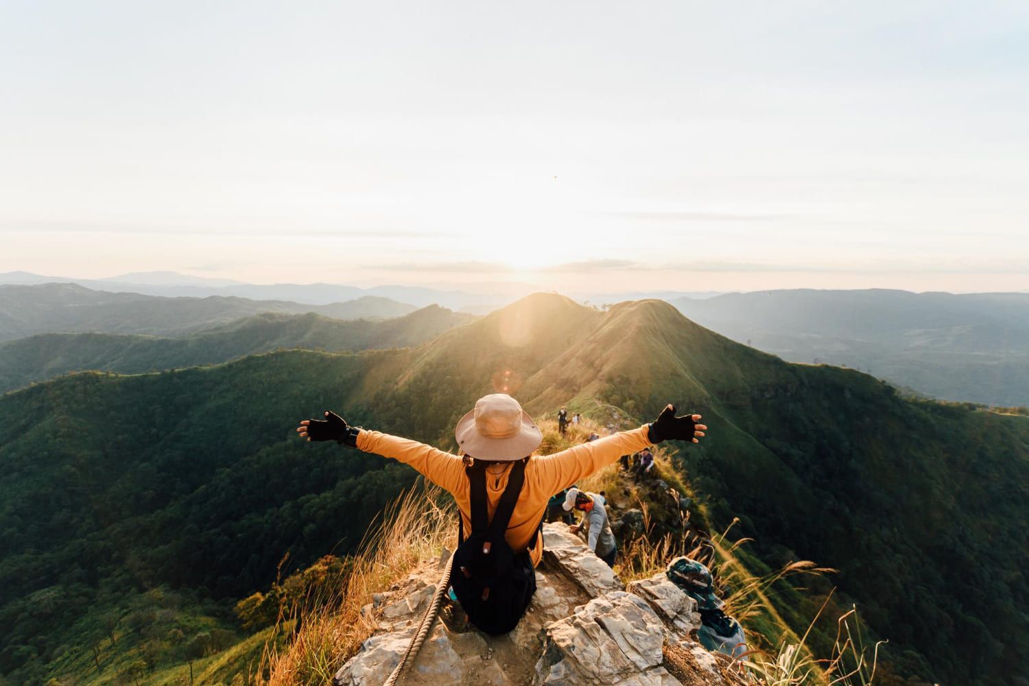 Mulher com os braços estendidos no pico da montanha, observando a paisagem verde ao pôr do sol.